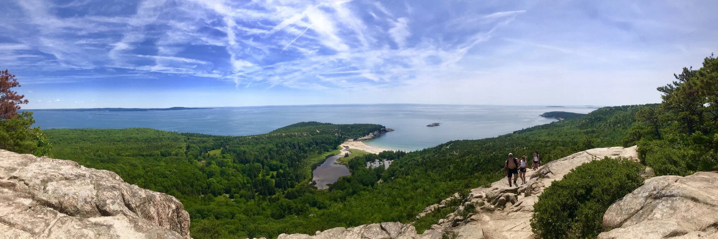 Scenic view from a hilltop overlooking a lush green forest, a sandy beach, a small bay, and the ocean with a partly cloudy sky and three hikers walking on a rocky trail.