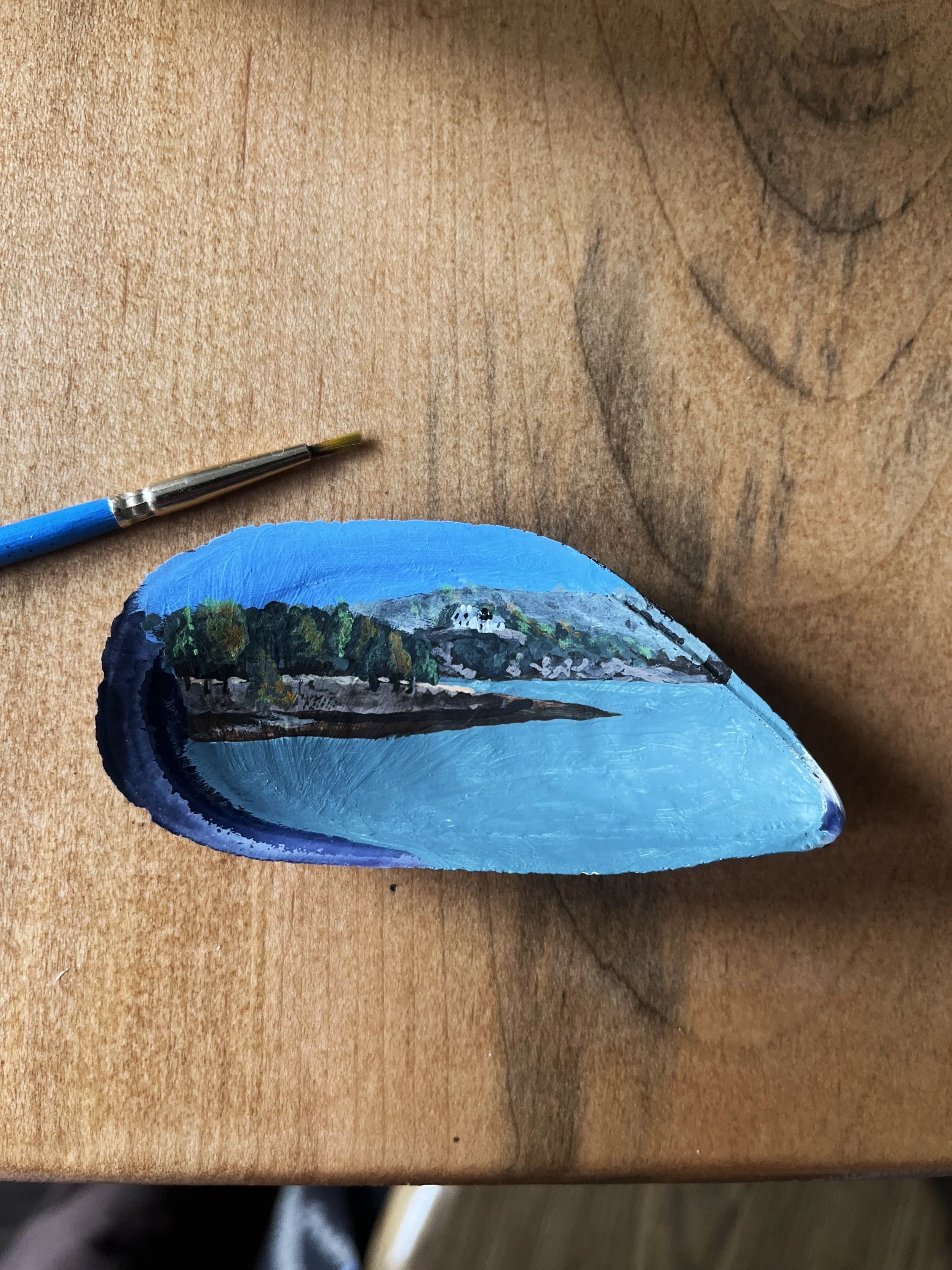A small painted mussel shell with a landscape scene featuring a blue sky, green trees, and a hill, placed on a wooden surface.