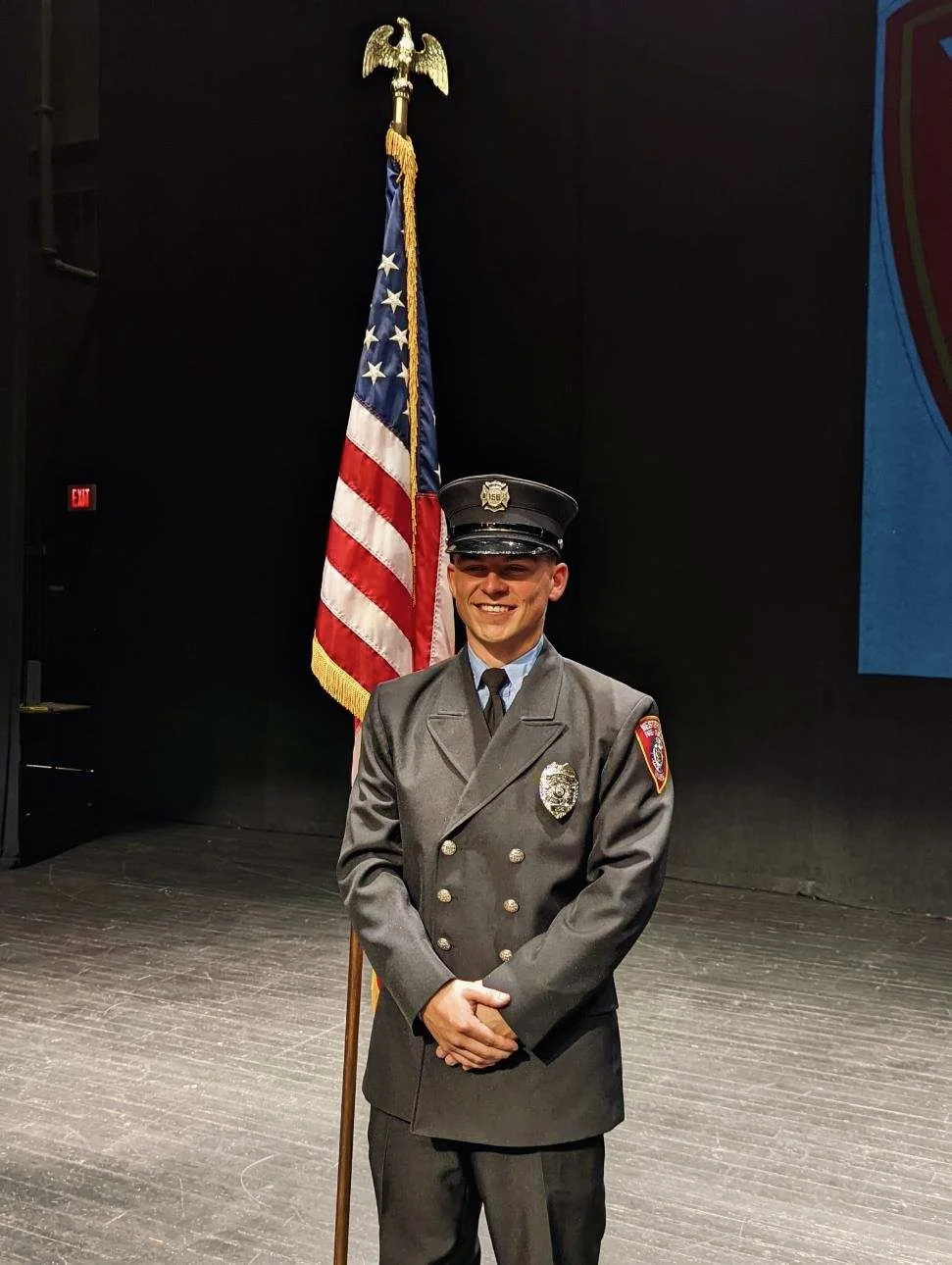 A smiling firefighter in uniform stands on a stage in front of an American flag, with a dark background.