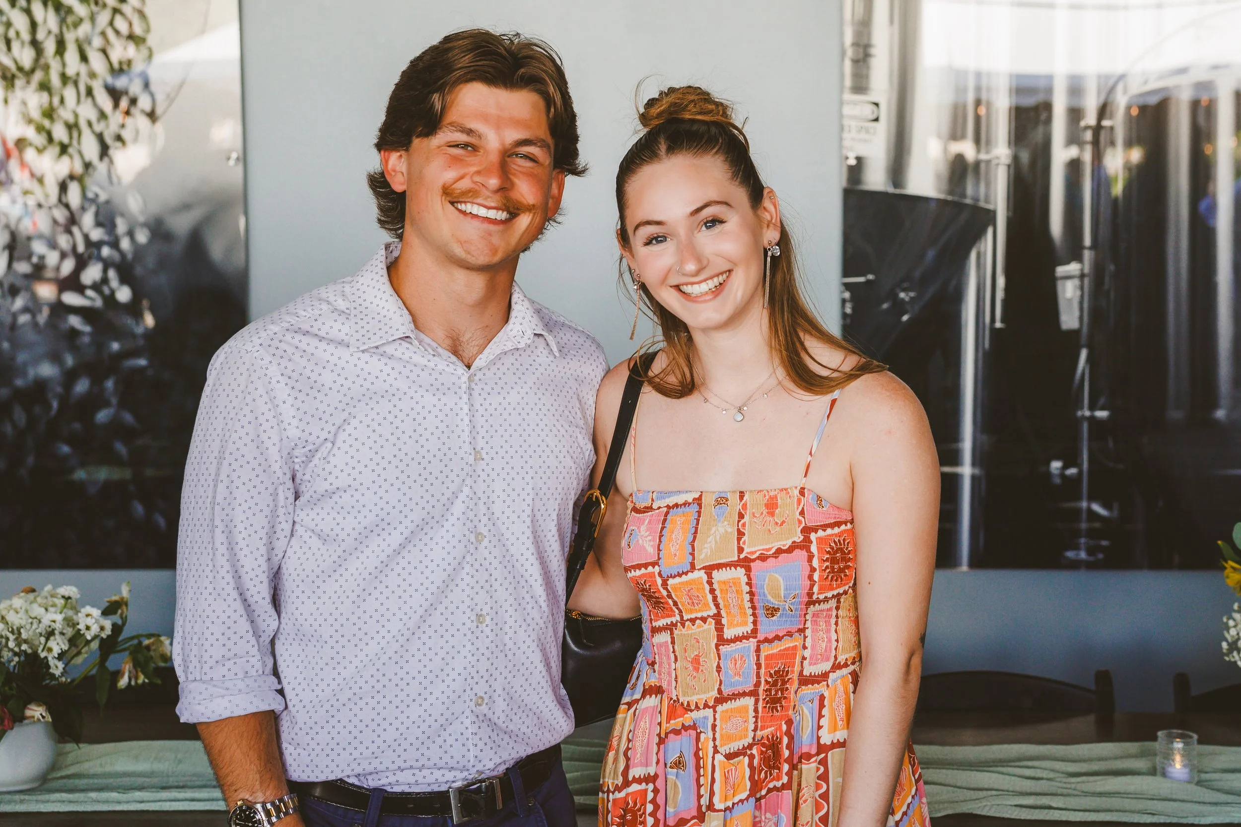 A smiling man wearing a white patterned shirt and a smiling woman wearing a colorful dress with a black purse standing close together indoors, with plants and artwork behind them.