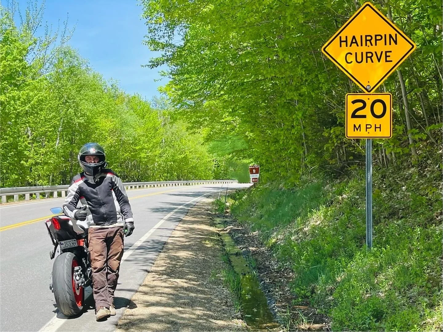 A motorcyclist standing on the side of a curved, two-lane road in a forested area under a blue sky with green trees. There are road signs indicating a hairpin curve and a speed limit of 20 mph.