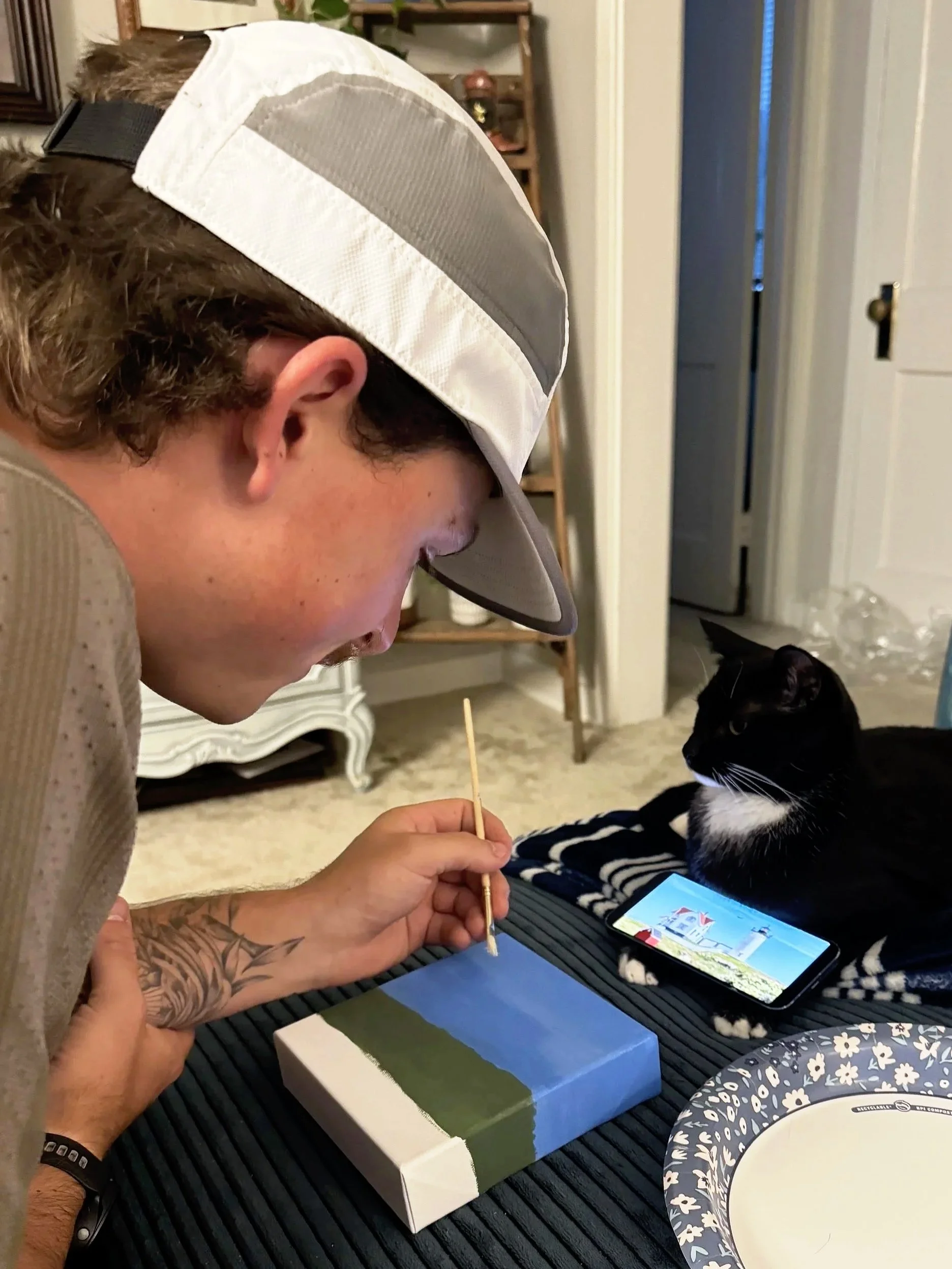 A person wearing a gray and white cap is painting a canvas with landscape scenery, using a fine brush, next to a black and white cat lying on a striped blanket. There is a smartphone displaying a landscape image and a decorative paper plate on the table.