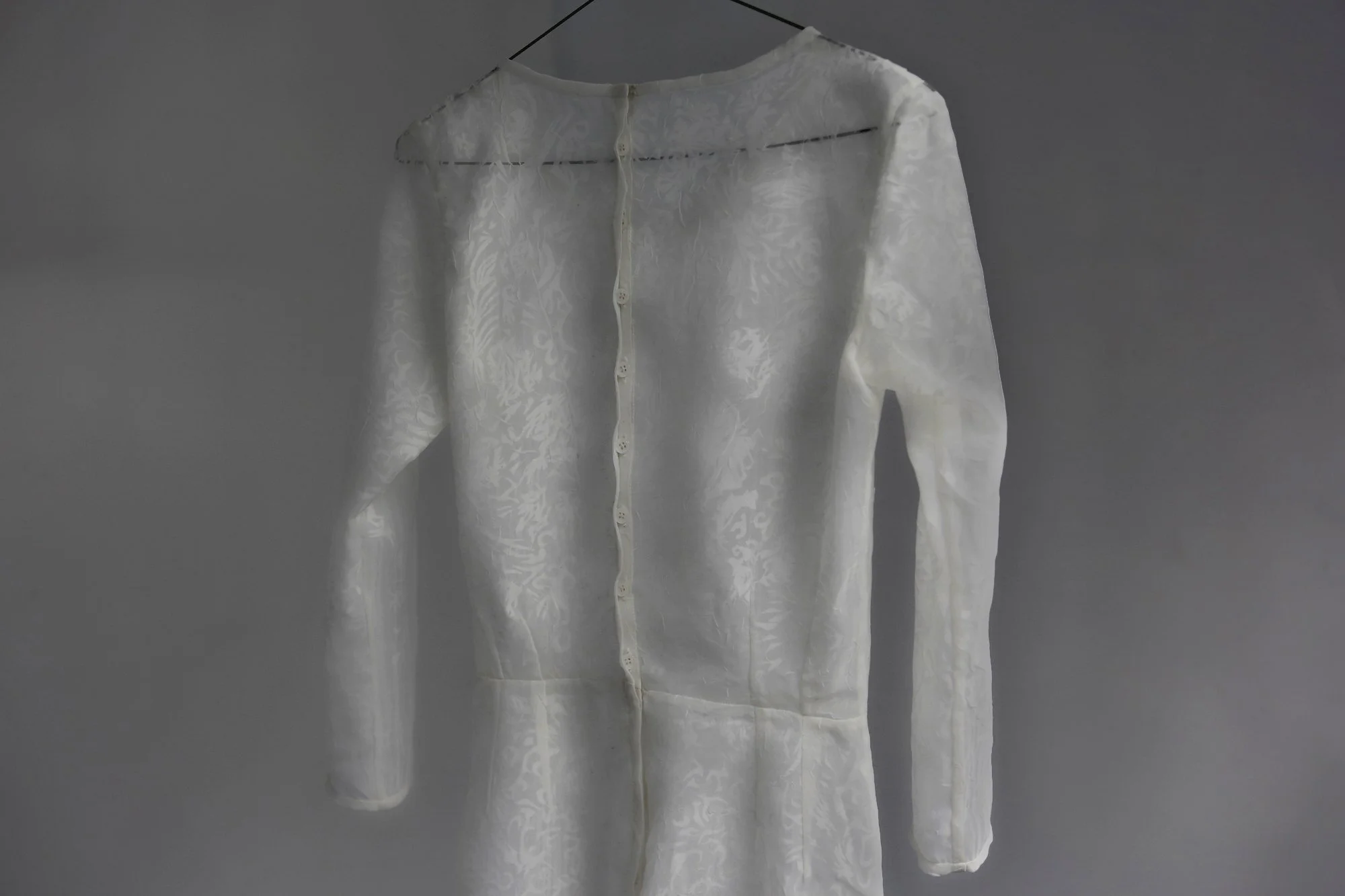 A white, long-sleeved, sheer button-up suit with a floral pattern that resembles an ancient chinese fabric pattern, hanging on a black wire hanger against a gray background.