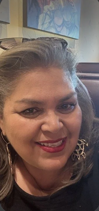 Close-up of a smiling woman with light skin, shoulder-length wavy hair, red lipstick, and large earrings, inside a restaurant with artwork on the wall in the background.