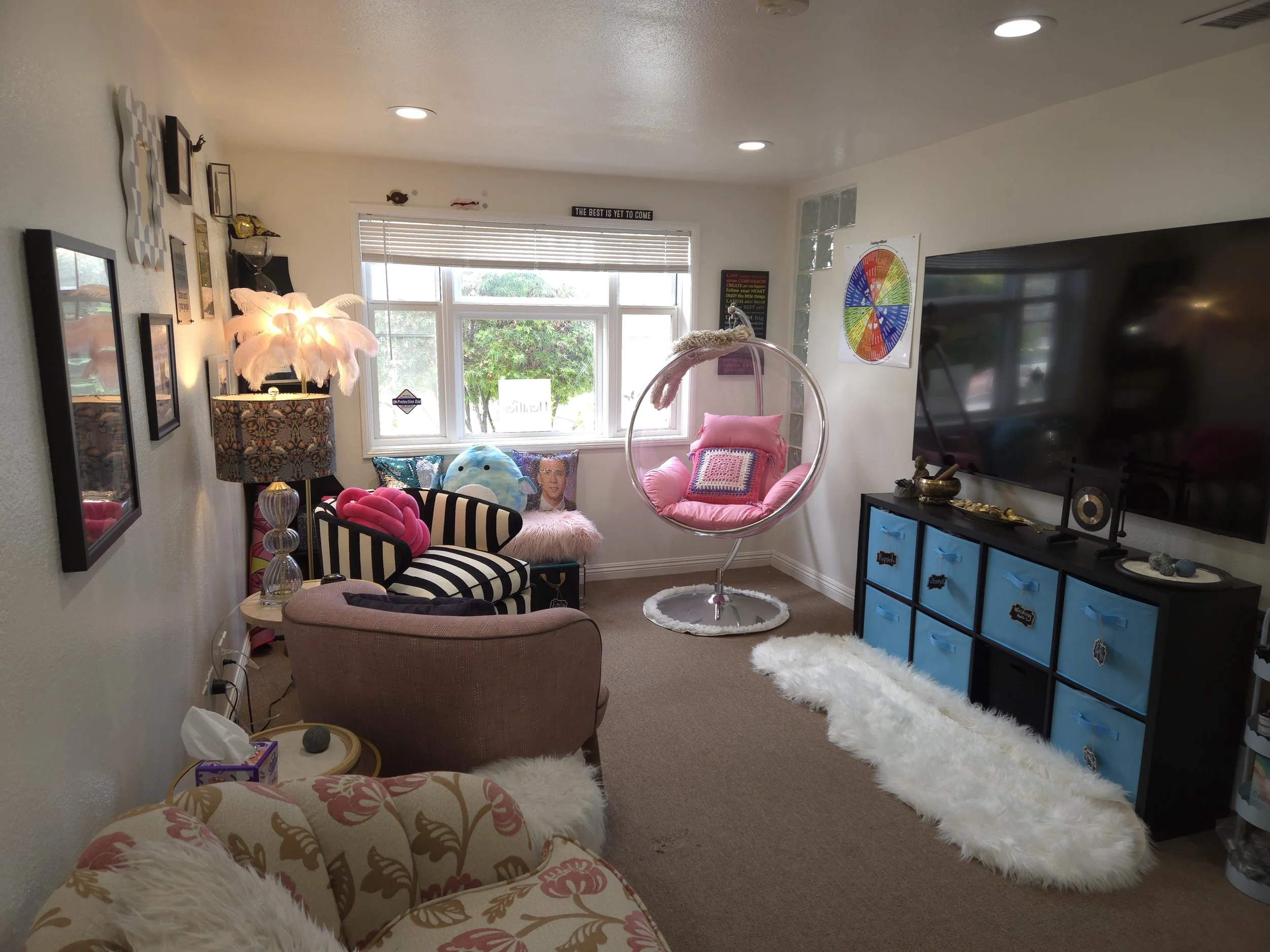 Living room with various chairs, a large TV, colorful decorations, a hanging bubble chair, and a window with greenery outside.