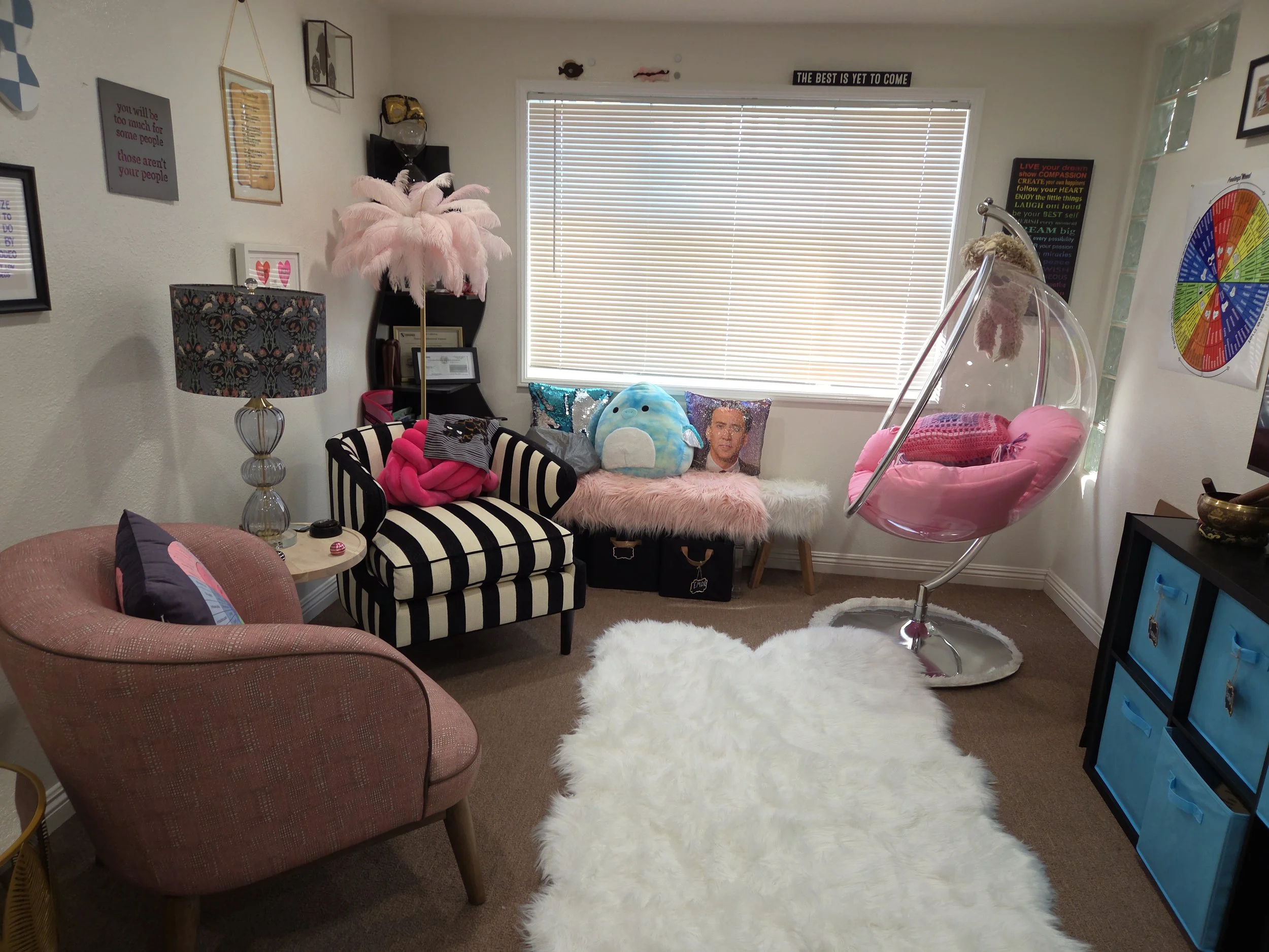 A cozy room decorated with colorful cushions, plush toys, and decorative wall art. There is a pink hanging chair, a fluffy white rug, and a window with blinds. The room has a mix of modern and playful decor.