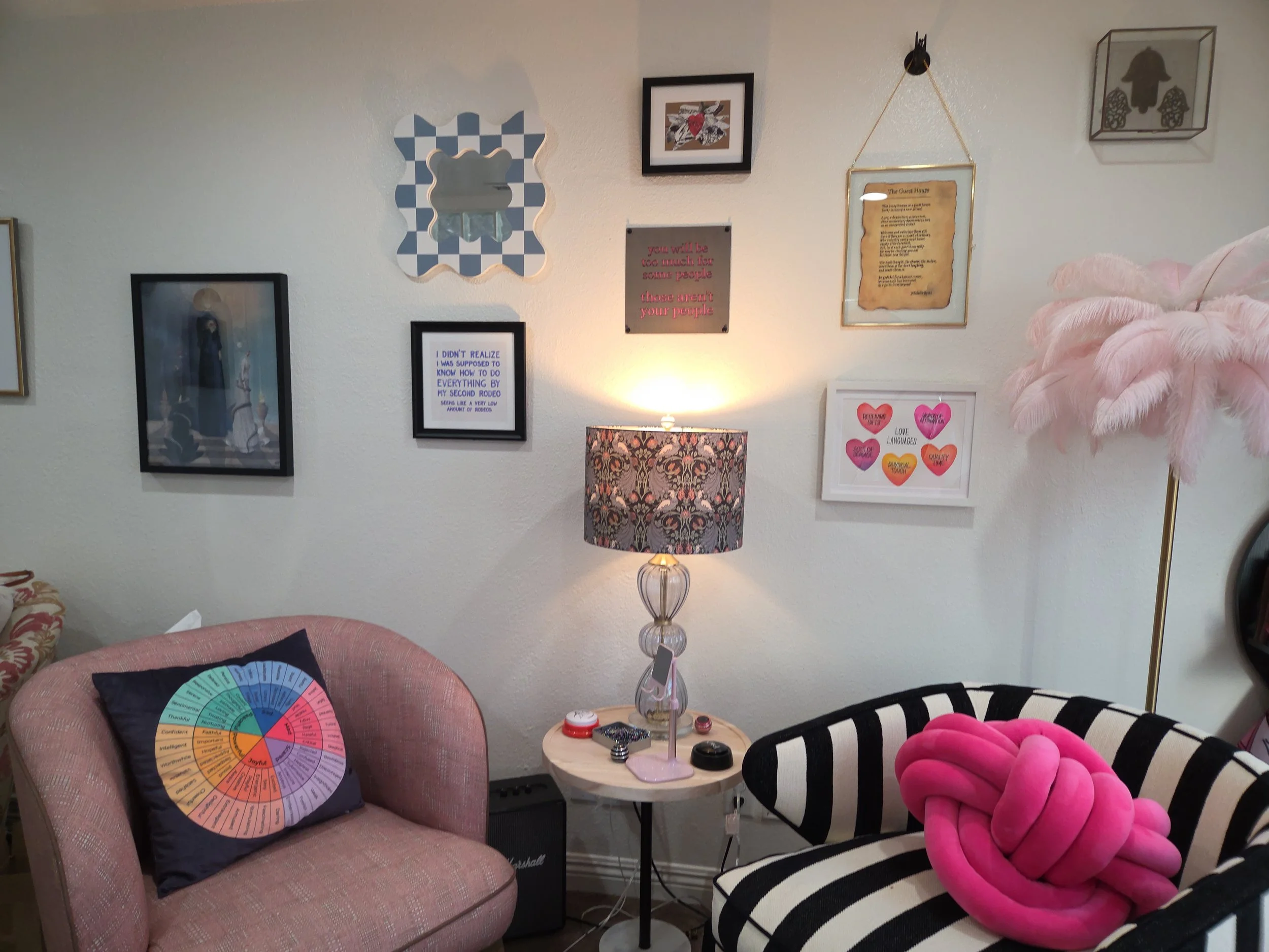 Living room with wall decorated with various framed art and quotes, pink upholstered armchair with colorful round art pillow, striped black and white chair with pink knotted cushion, side table with a floral lamp, and pink feather floor lamp.