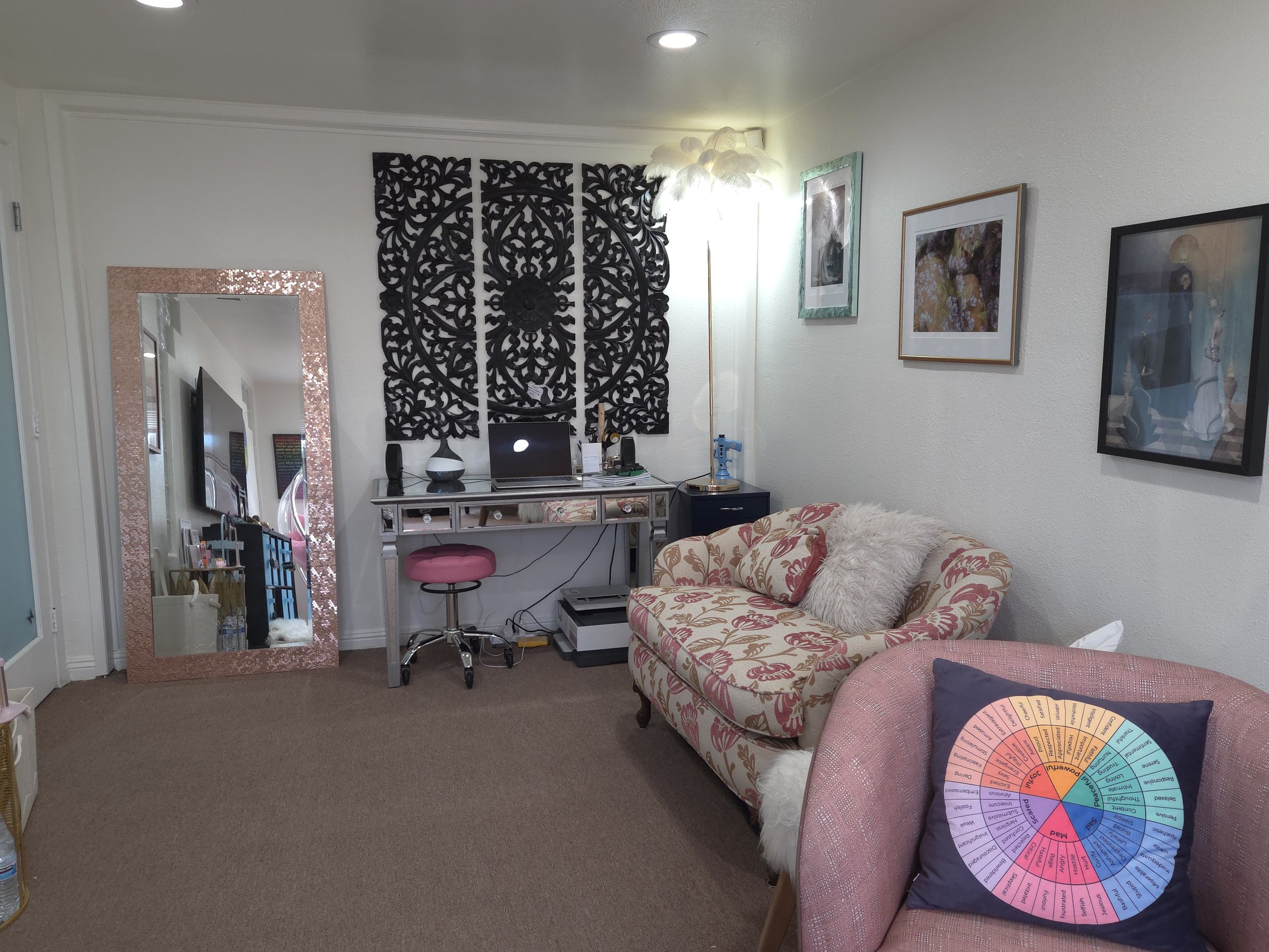 Cozy living room with a pink armchair, decorative pillows, wall art, a mirror, a desk with a laptop, chair, and a large ornate black wall hanging.