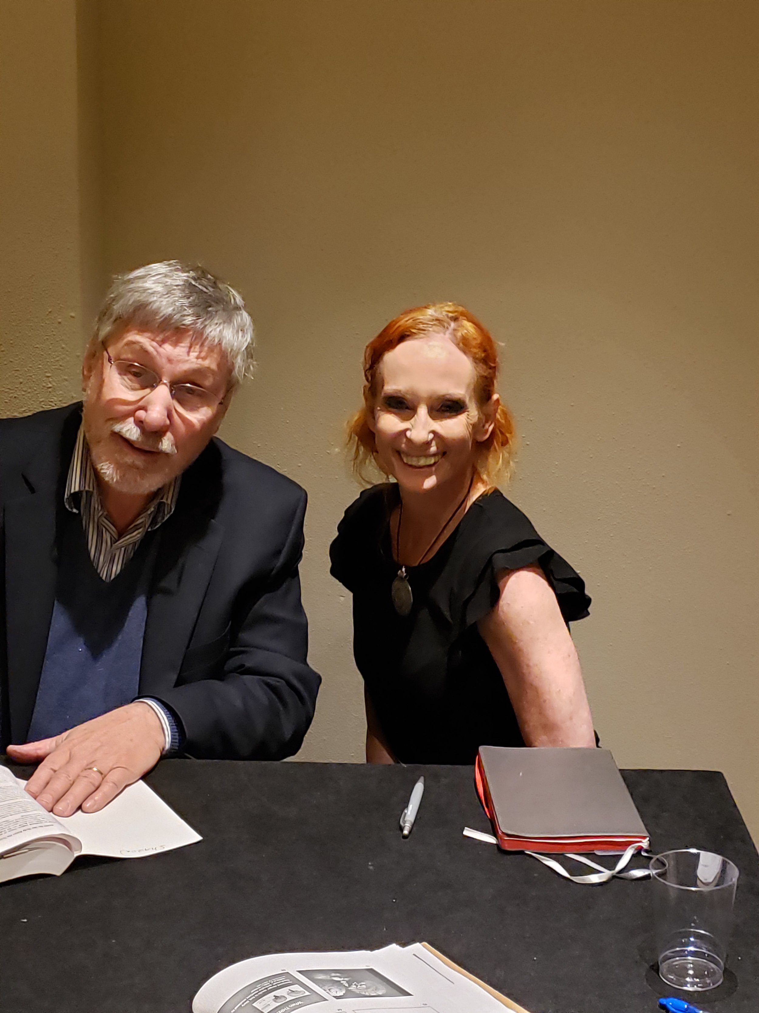 Two people, a man with glasses and gray hair and a woman with red hair, sitting at a table with books, notes, a pen, a glass of water, and other items, in a room with beige walls.
