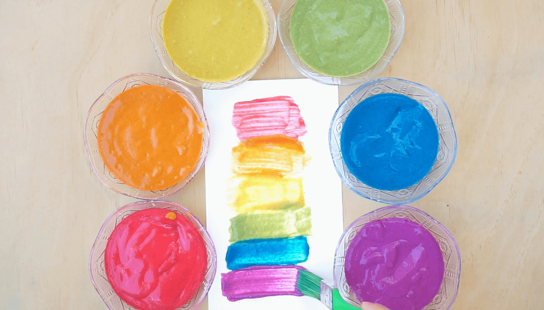 Six bowls of colorful slime arranged in a circle around a white sheet with a rainbow-colored swatch painted on it, on a wooden surface.