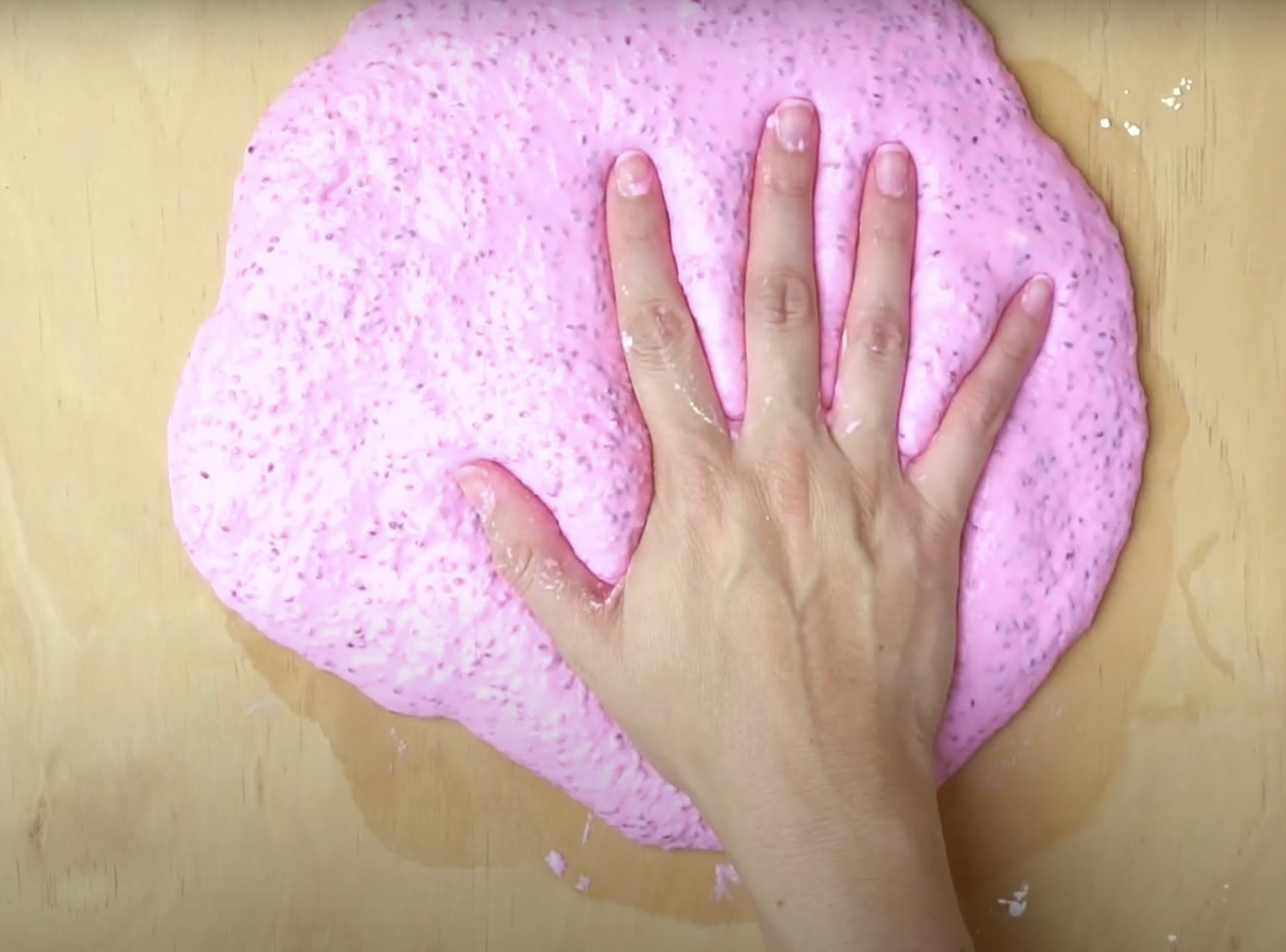 A hand pressing down on a bright pink slime with a speckled texture on a wooden surface.