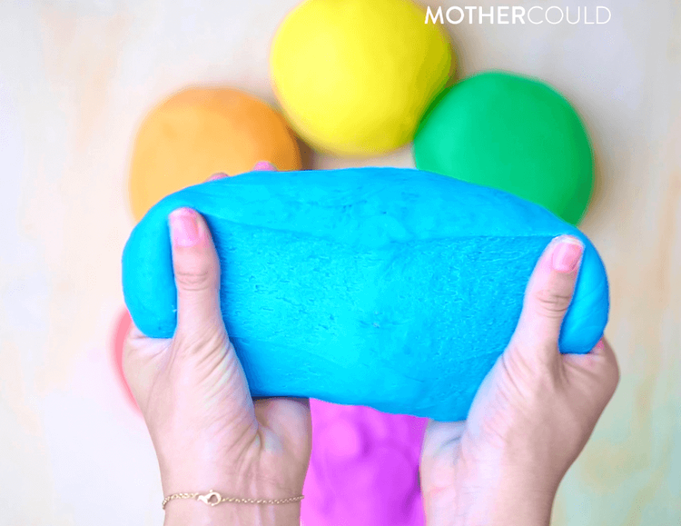 Person squeezing a blue balloon in front of three blurred balloons in yellow, orange, and green, with the text 'Mother Could' in the top right corner.