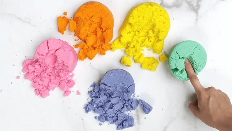 Five colorful chalk powders in pink, orange, yellow, blue, and green on a white surface. A hand with a finger touching the green chalk powder.