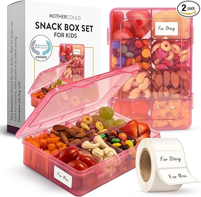 Pink snack box with multiple compartments filled with assorted nuts, dried fruit, and candies. Includes a roll of labels for personalization, with one label saying "For Stacey" and another "For Mom." The packaging indicates it is a kids' snack box set by Mother Could, and shows a two-pack of the snack containers.