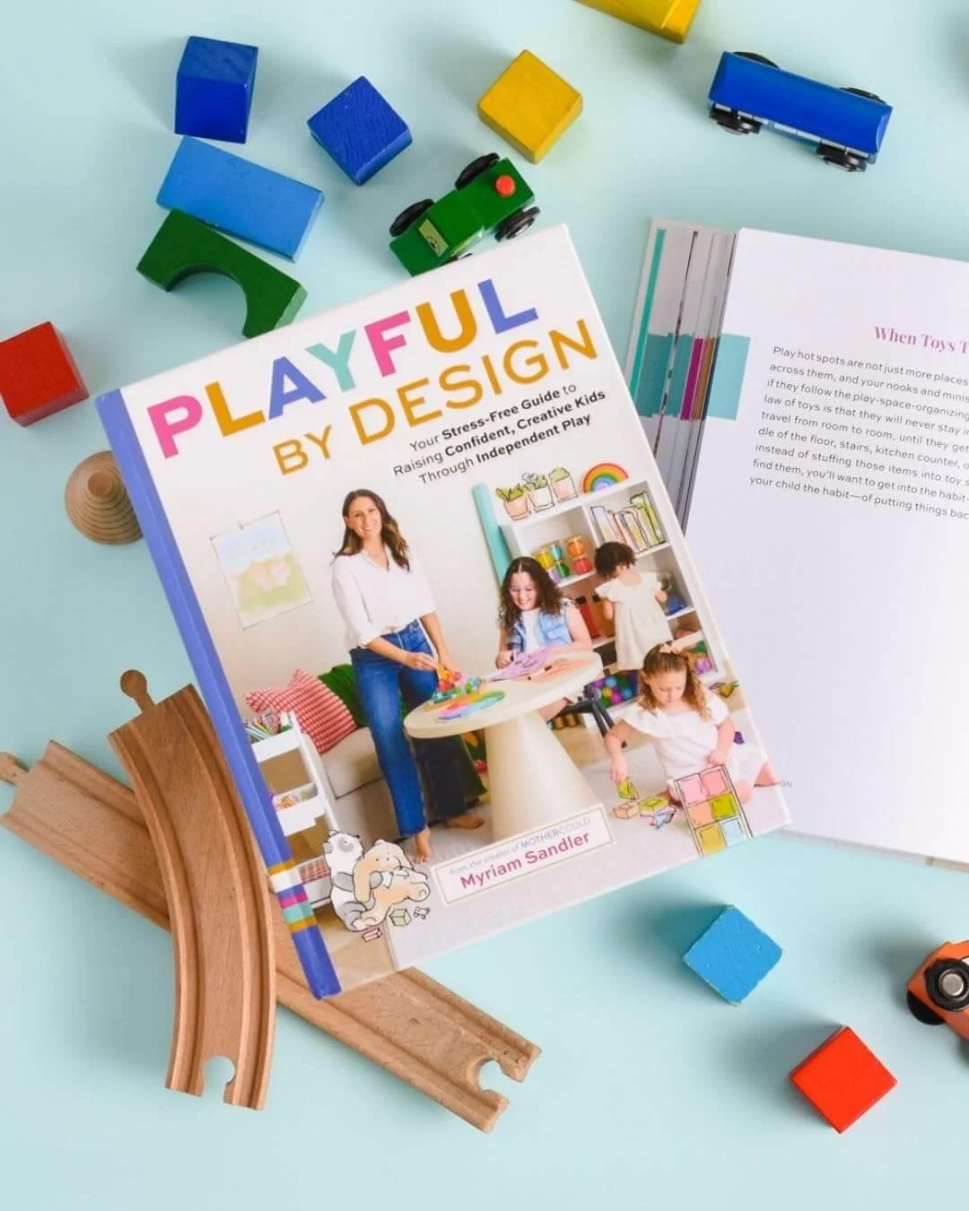 A book titled 'Playful by Design' placed on a light blue surface, surrounded by colorful toy blocks, a wooden train track, and other small toys. The book features a woman and children on the cover playing.