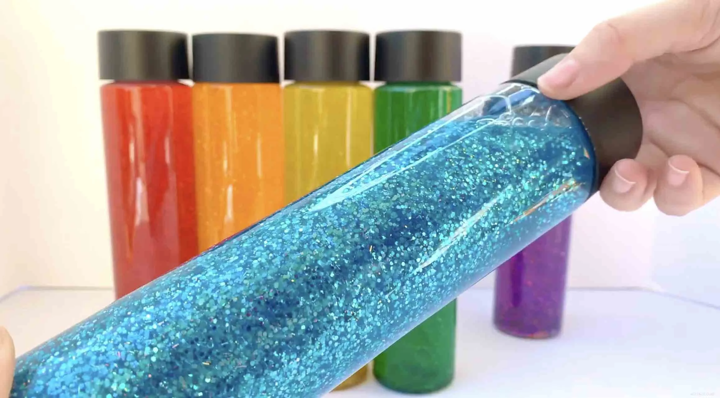 A hand holding a clear plastic tube filled with blue glitter, with colorful glitter bottles in the background.