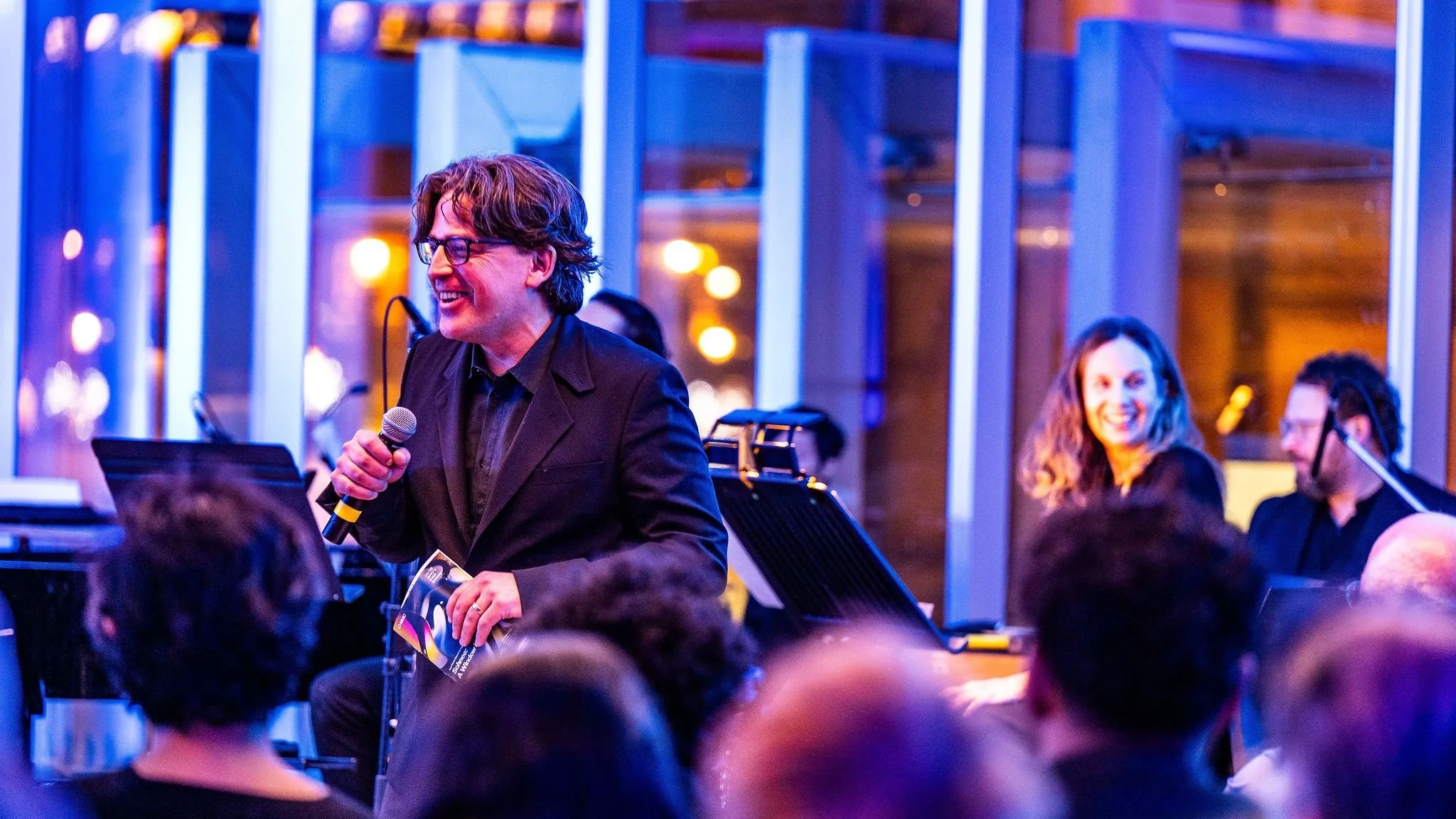 A man in glasses holding a microphone and a booklet, smiling while speaking at an event. Several people are seated around him, some of whom are musicians with instruments.