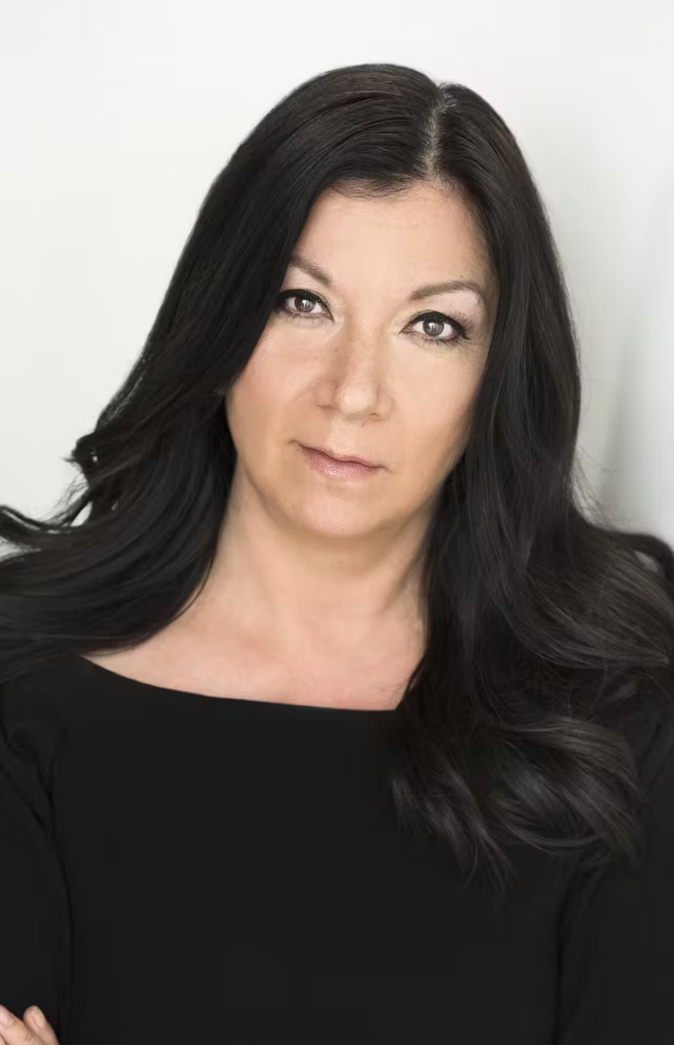 A woman with long black hair, wearing a black top, looking into the camera with a neutral expression against a plain white background.