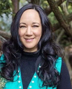 A woman with long dark wavy hair smiling outdoors with trees in the background.