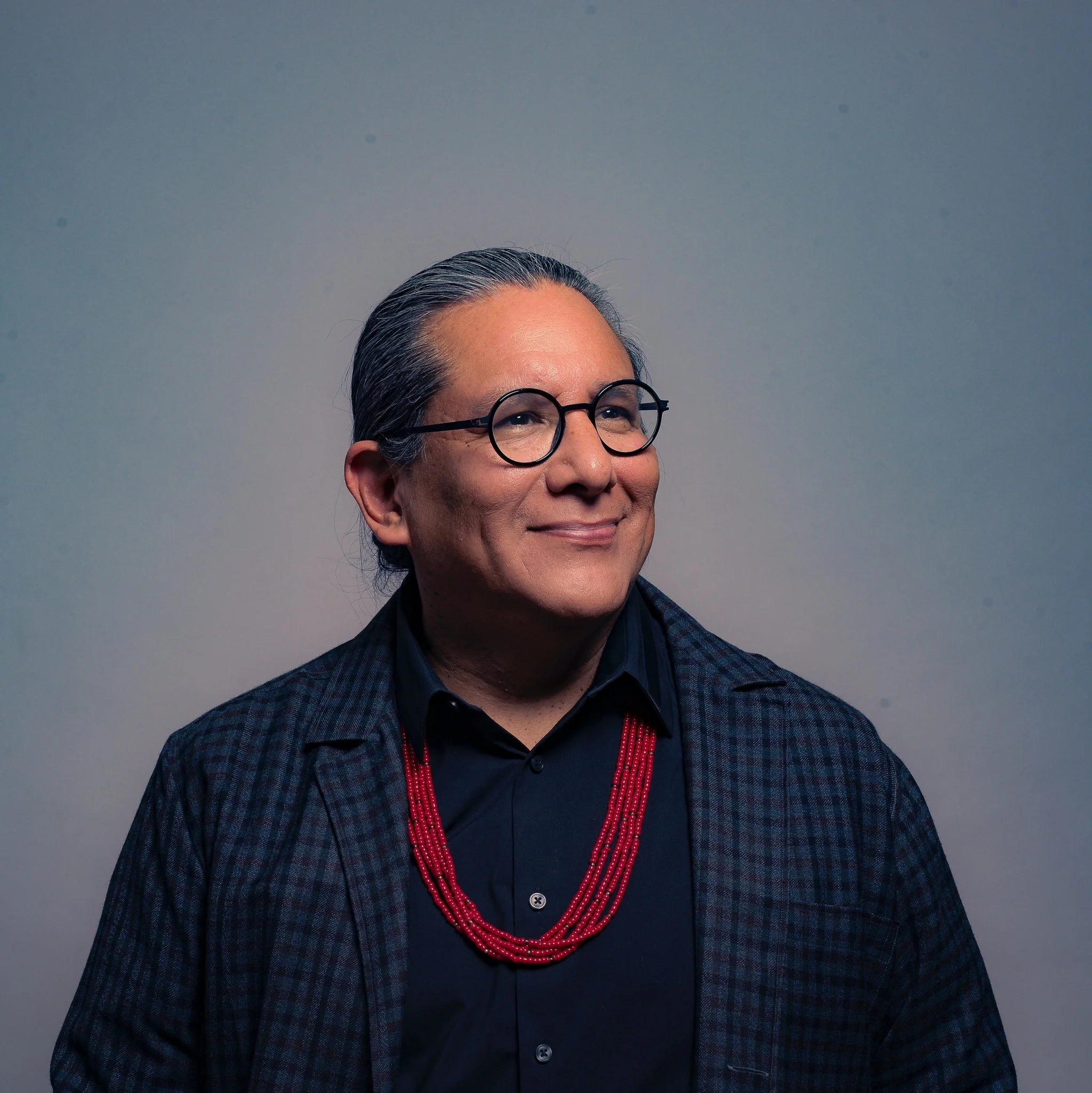 Portrait of a middle-aged man with gray hair, round glasses, smiling, wearing a dark shirt, red beaded necklace, and checkered blazer, against a plain background.