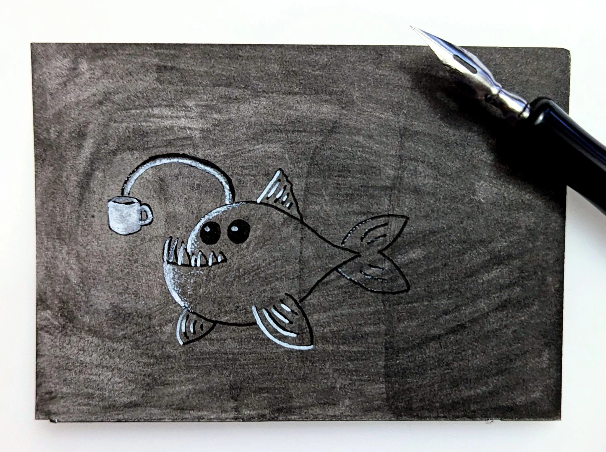 Drawing of a cartoon anglerfish with big black eyes, fins, and a tail, using a coffee as a lure. the fountain pen used to draw the fish is in the picture.