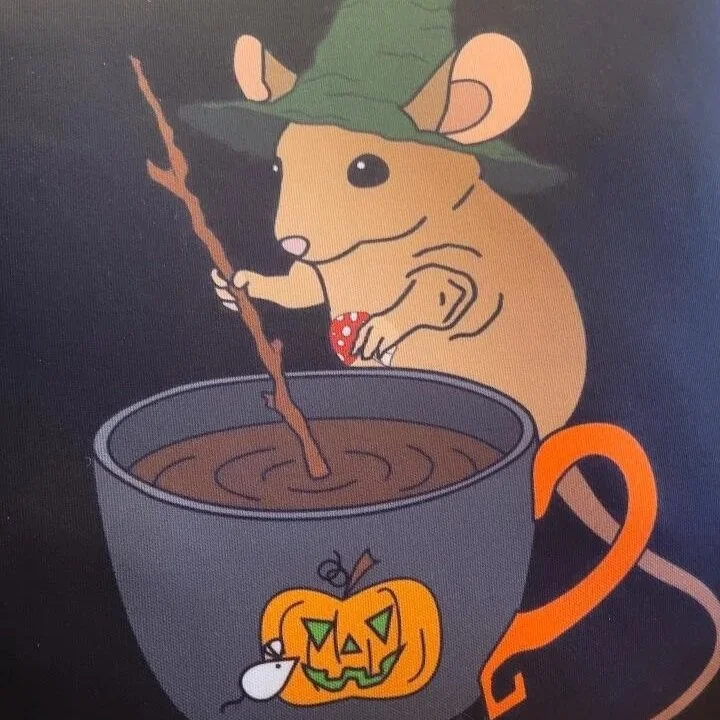 A cartoon mouse dressed as a witch, wearing a green hat, stirring a hot chocolate in a mug with a Halloween pumpkin on it.