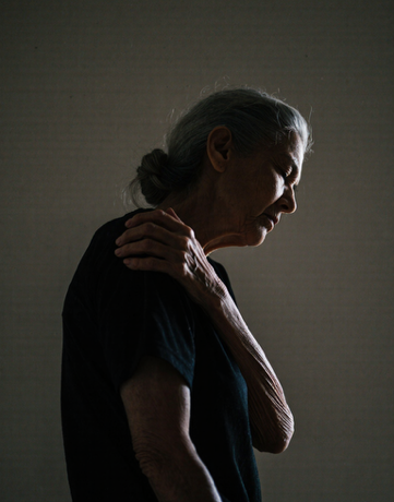 Side profile of an elderly woman with gray hair, touching her neck in pain, against a plain background.