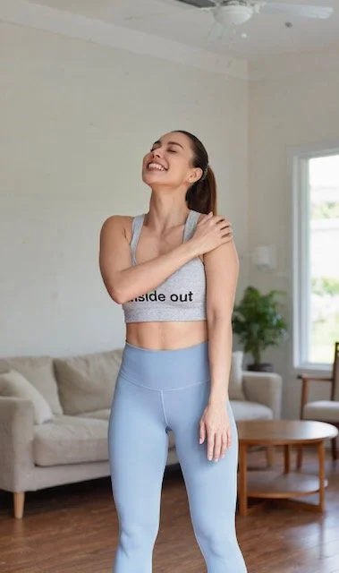 A woman smiling and stretching in a living room, wearing a gray crop top with 'insideout' written on it and light blue leggings.