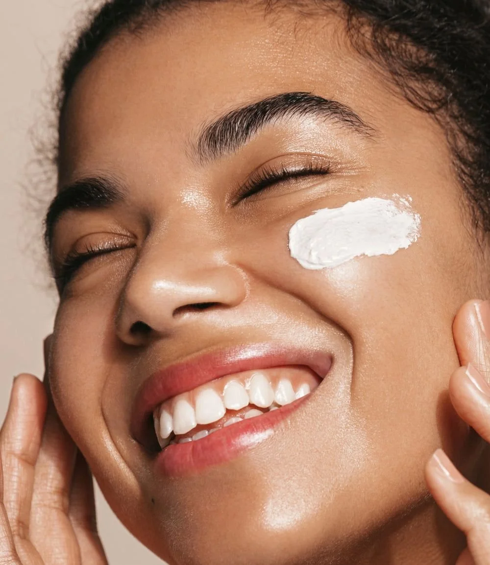 A woman smiling with a white cream applied under her eye, touching her face gently.