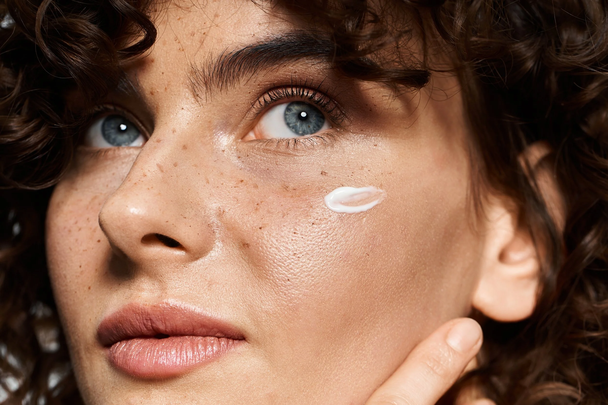 Close-up of a woman with curly hair applying a white cream to her cheek.