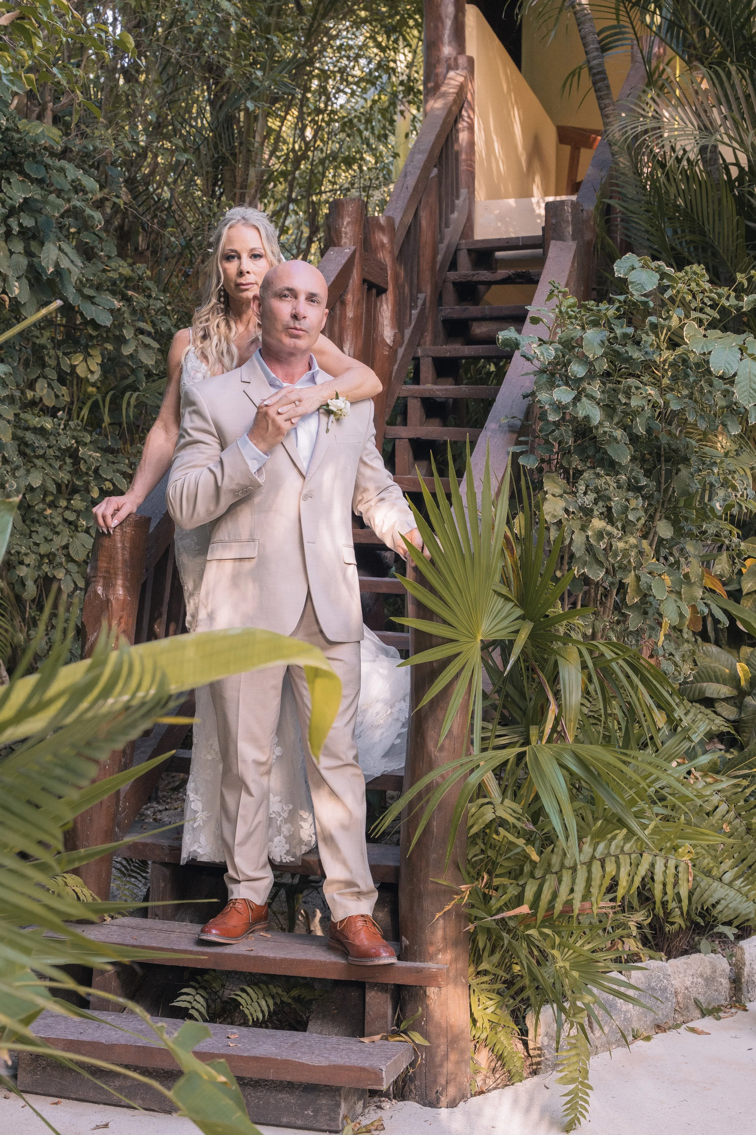 A couple in wedding attire standing on a wooden staircase amidst lush greenery, with the woman behind and over the man, both serious expressions.