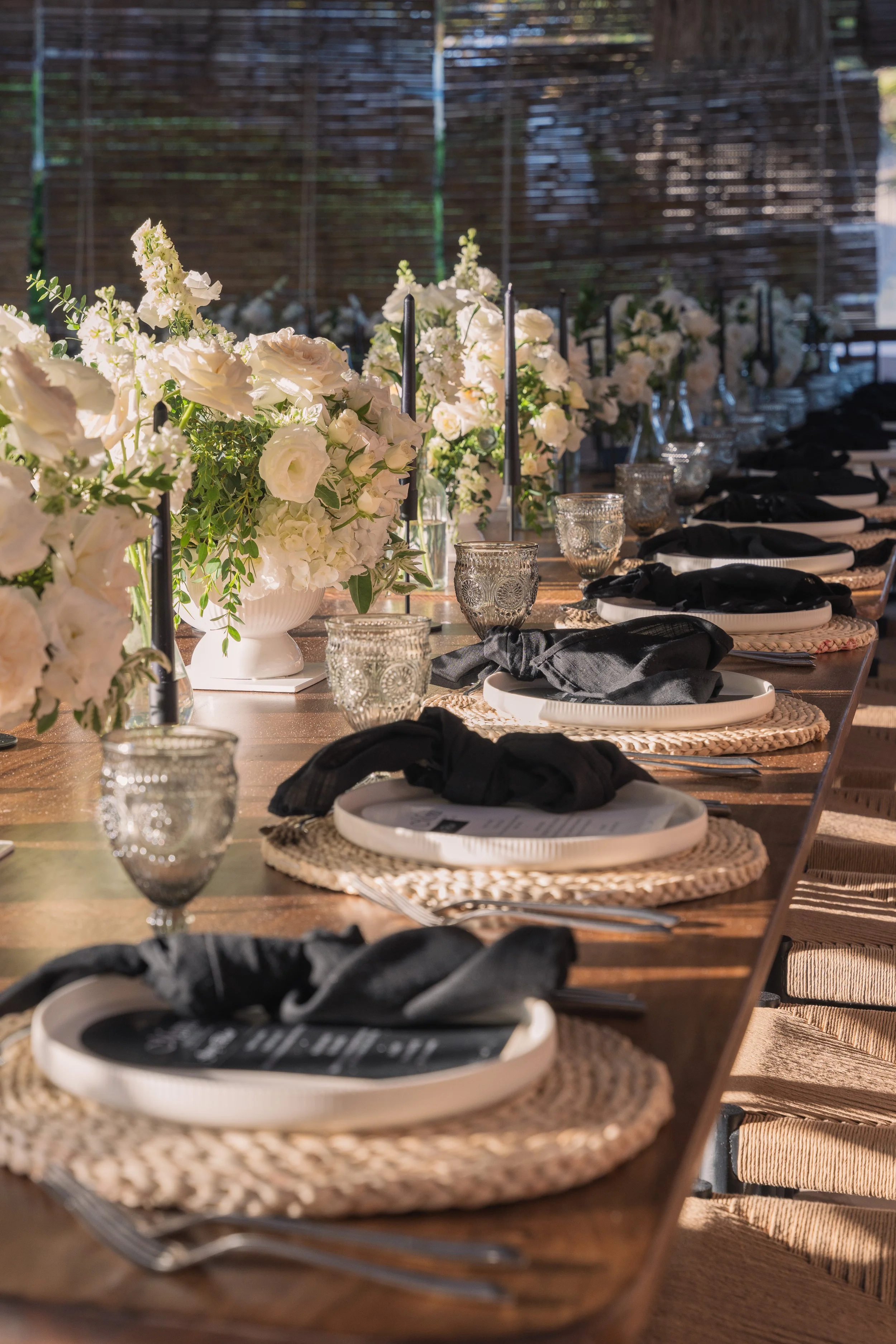 Wedding reception table with white floral arrangements, black napkins, glass goblets, and woven placemats.