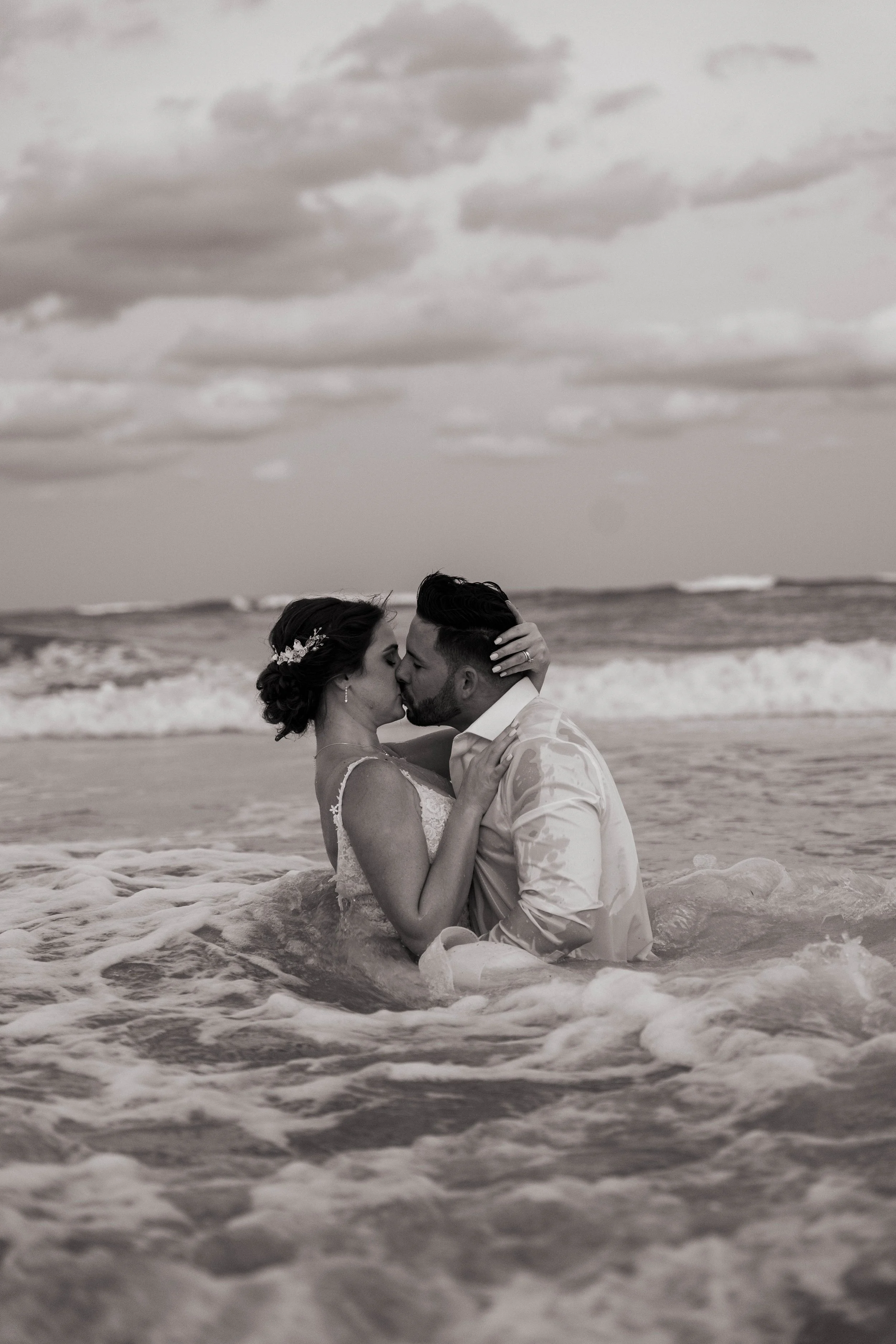 Elopement wedding. Couple kissing in the ocean, waves in foreground, cloudy sky in background
