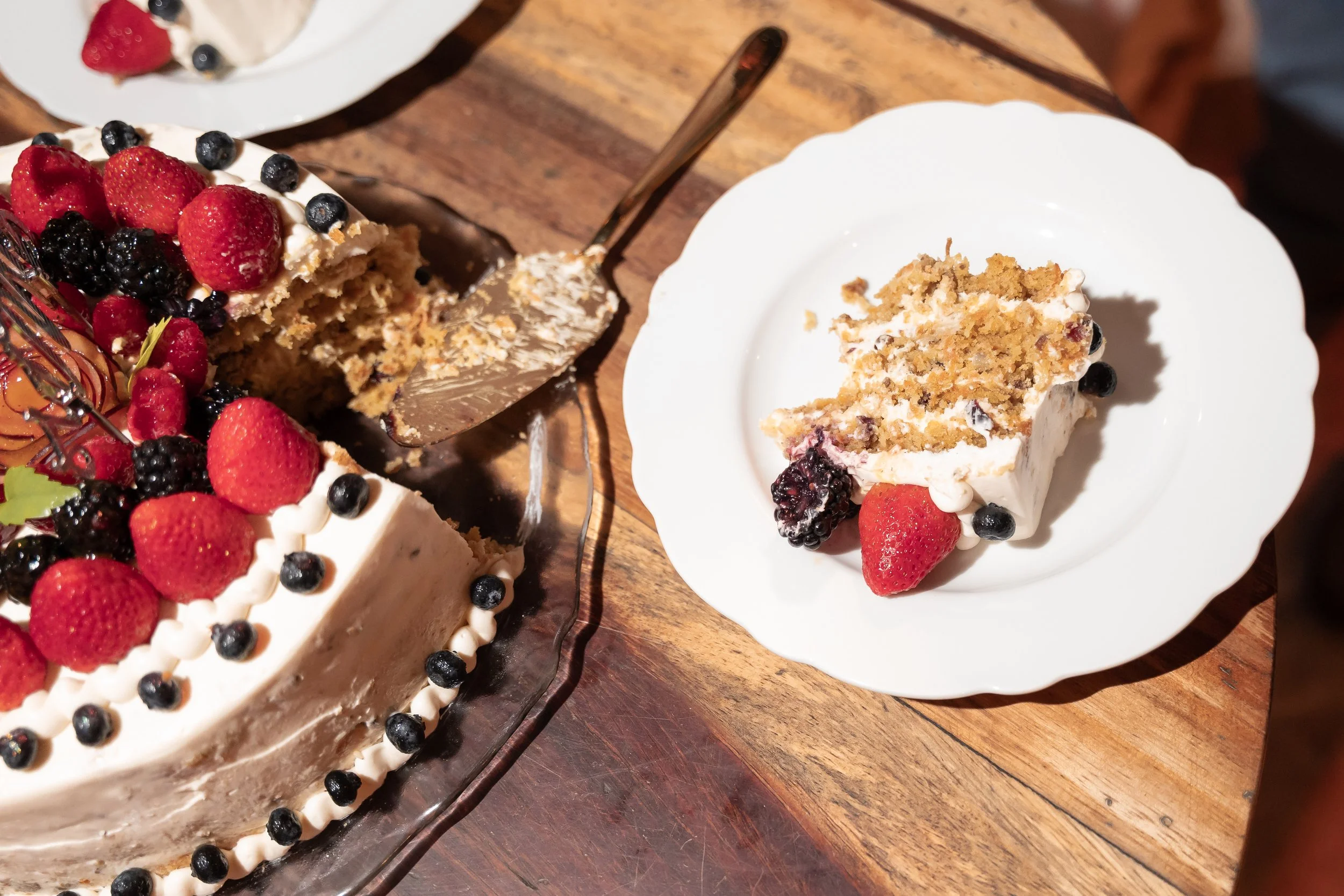 A slice of layered cake with white frosting, topped with assorted fresh berries including strawberries, blackberries, and blueberries, served on a white plate. A larger cake with similar toppings is partially visible in the background on a decorative