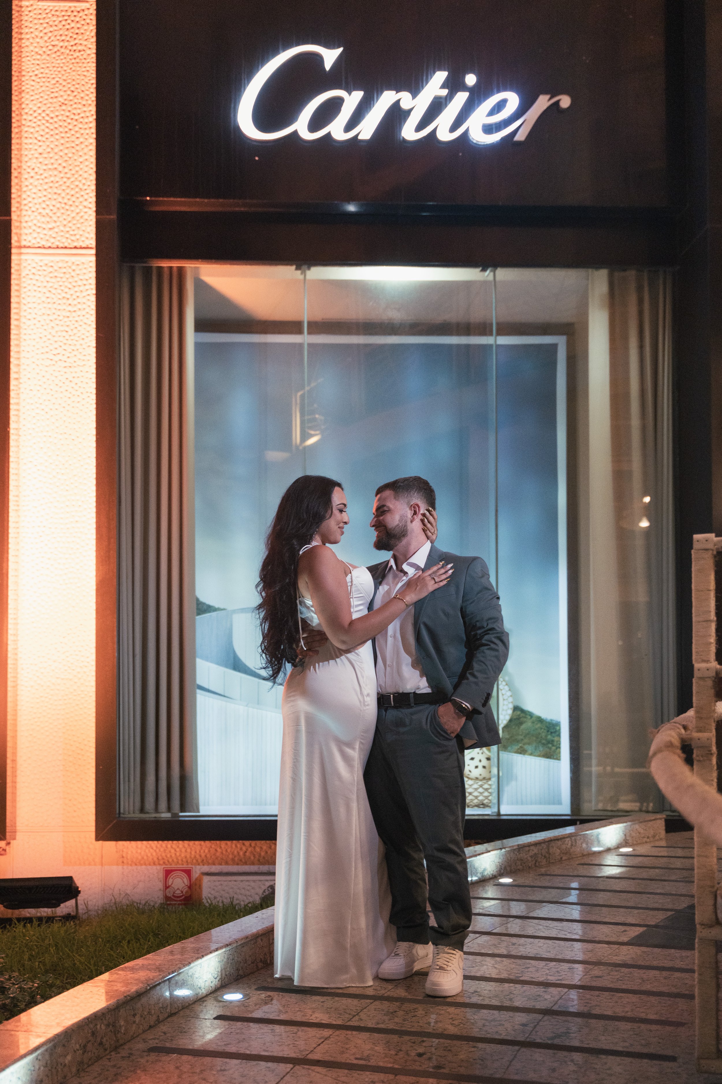 A couple standing close together outside a Cartier store at night, with the woman wearing a white dress and the man in a suit, sharing an intimate moment.