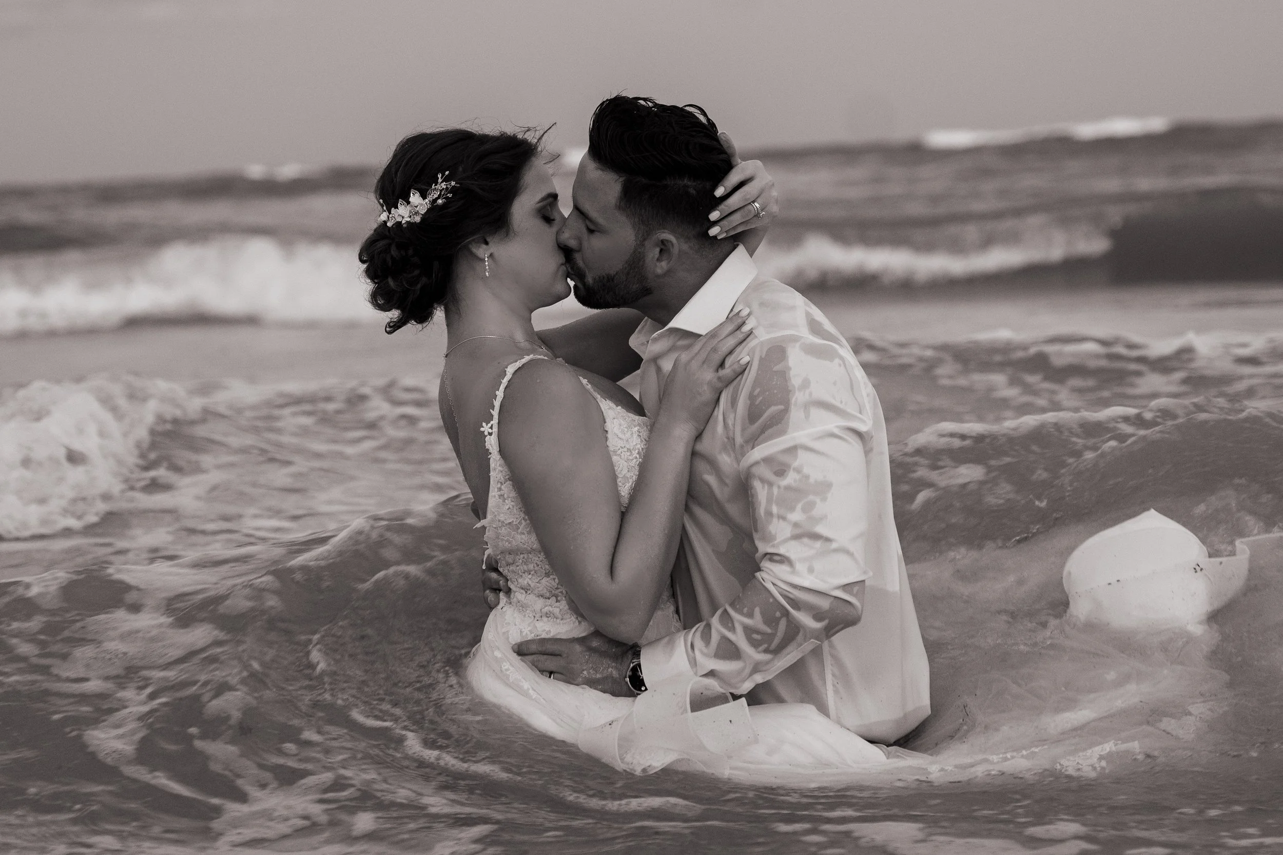 A couple in wedding attire sharing a kiss while standing in the ocean, with waves around them, during sunset.