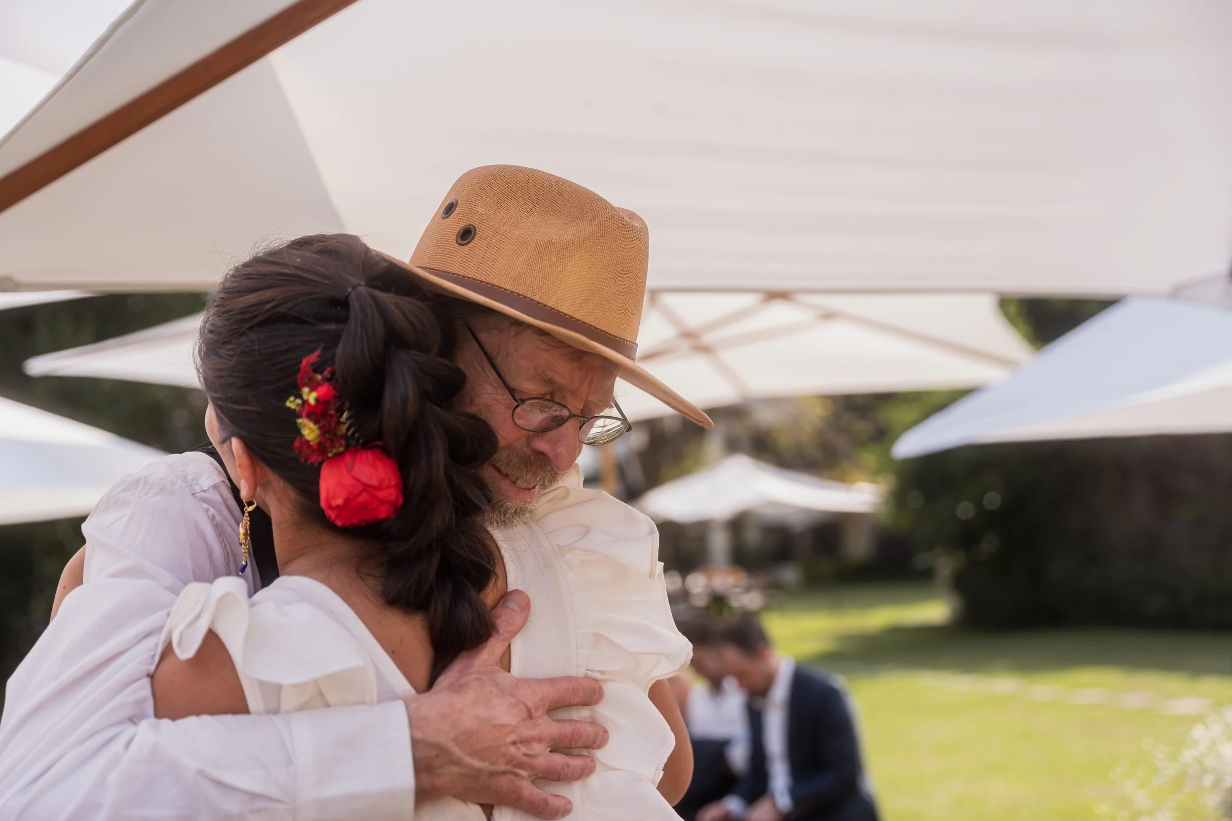 An elderly man wearing a tan hat and glasses hugging a woman with dark hair adorned with flowers, at an outdoor event under white umbrellas.