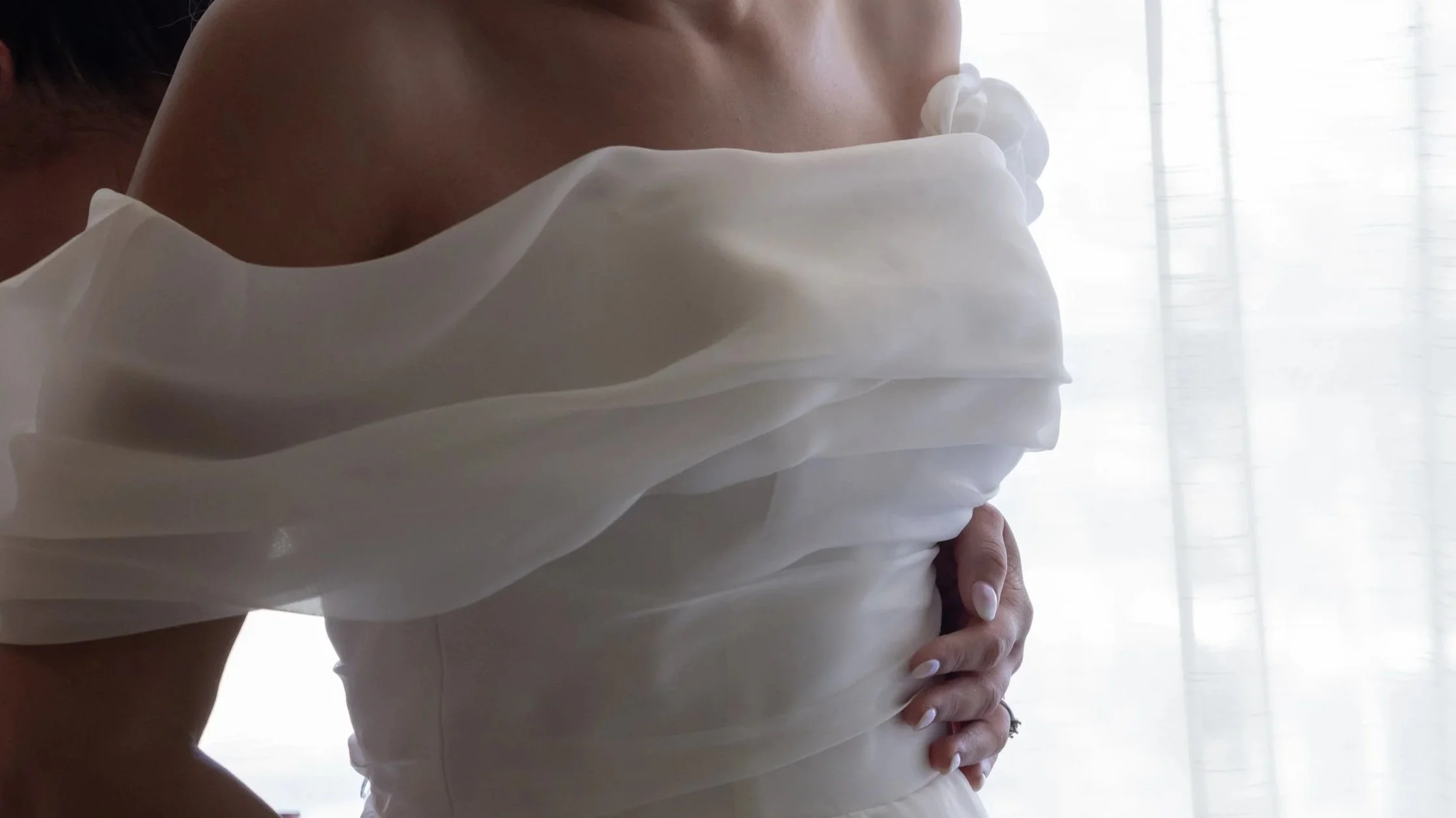 A woman in a white dress with sheer fabric on her shoulders, standing indoors near a light-colored curtain.