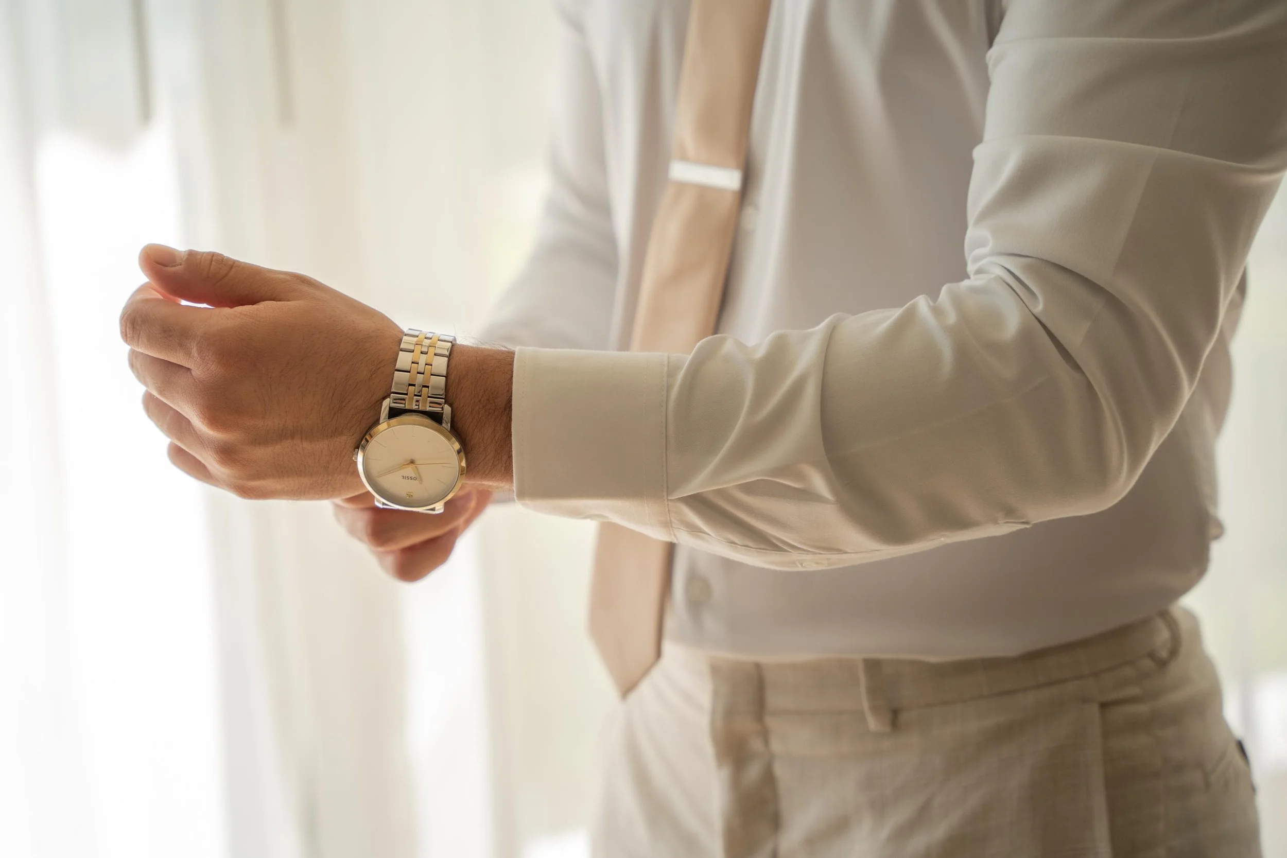 Close-up of a man wearing a beige and silver wristwatch, adjusting it with his other hand, dressed in a white shirt and light-colored pants, in a well-lit room.