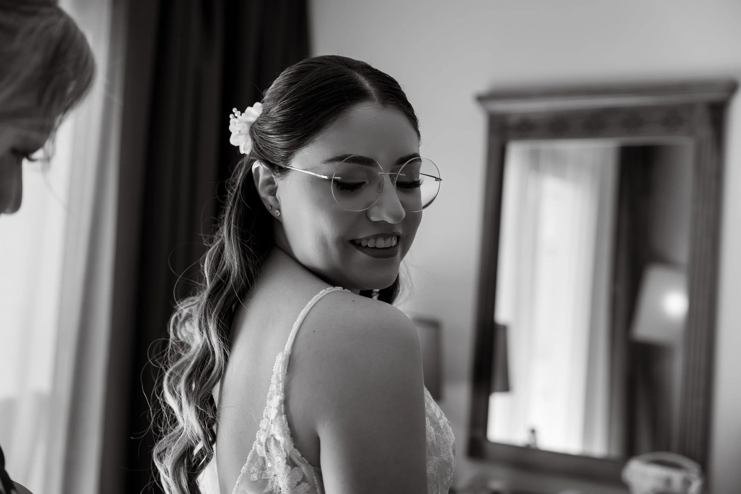 A woman smiling with her eyes closed, wearing glasses and a dress with lace straps, standing in front of a mirror in a room.