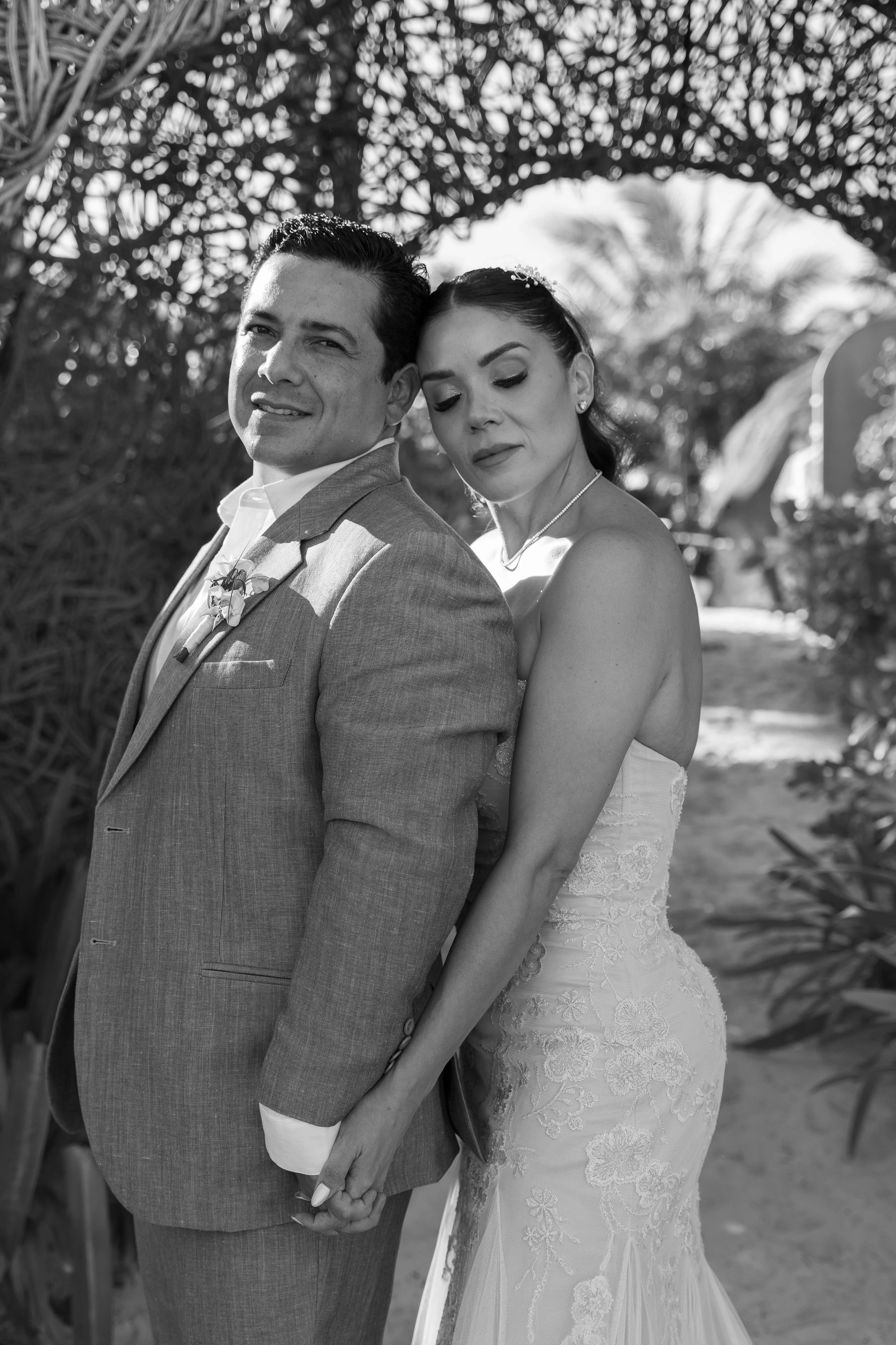 Black and white photo of a bride and groom holding hands, standing close together outdoors at Tulum with tropical plants and trees in the background. They are a couple from Mexico’s political and business elite.
