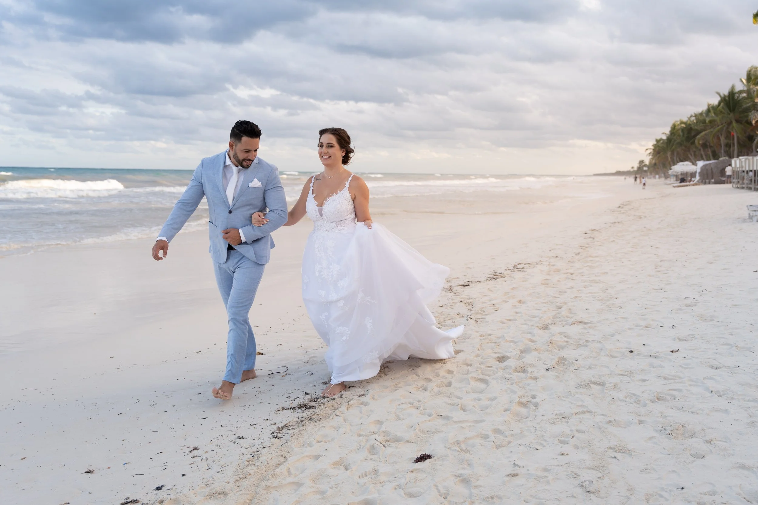 A bride and groom walking barefoot on the beach, holding hands and smiling, with palm trees and beach umbrellas in the background.