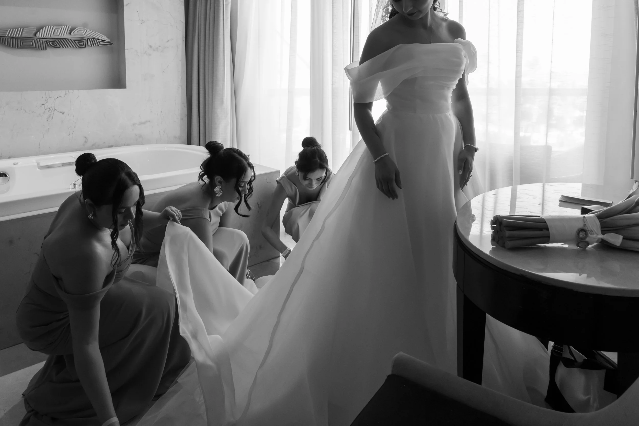 A bride in a wedding dress with four bridesmaids helping her with the dress in a hotel room.