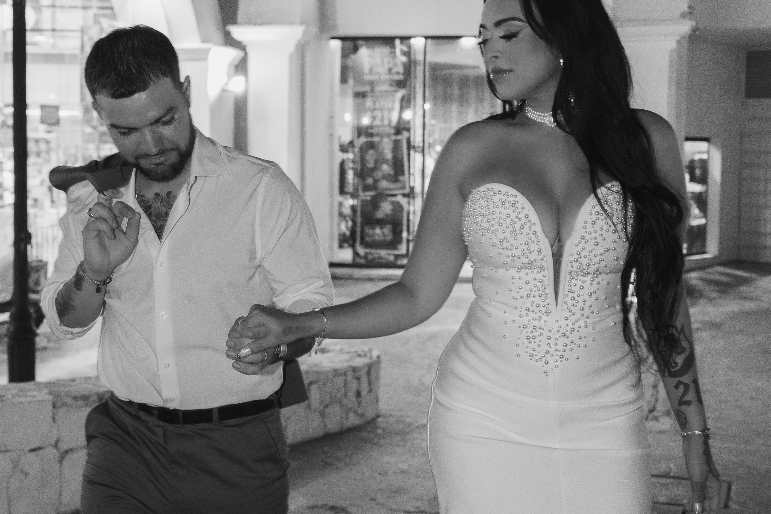 A man and a woman holding hands and walking outside at night. The woman is wearing a strapless dress with embellishments and has long dark hair. The man is dressed in a button-up shirt and pants, carrying a jacket over his shoulder.