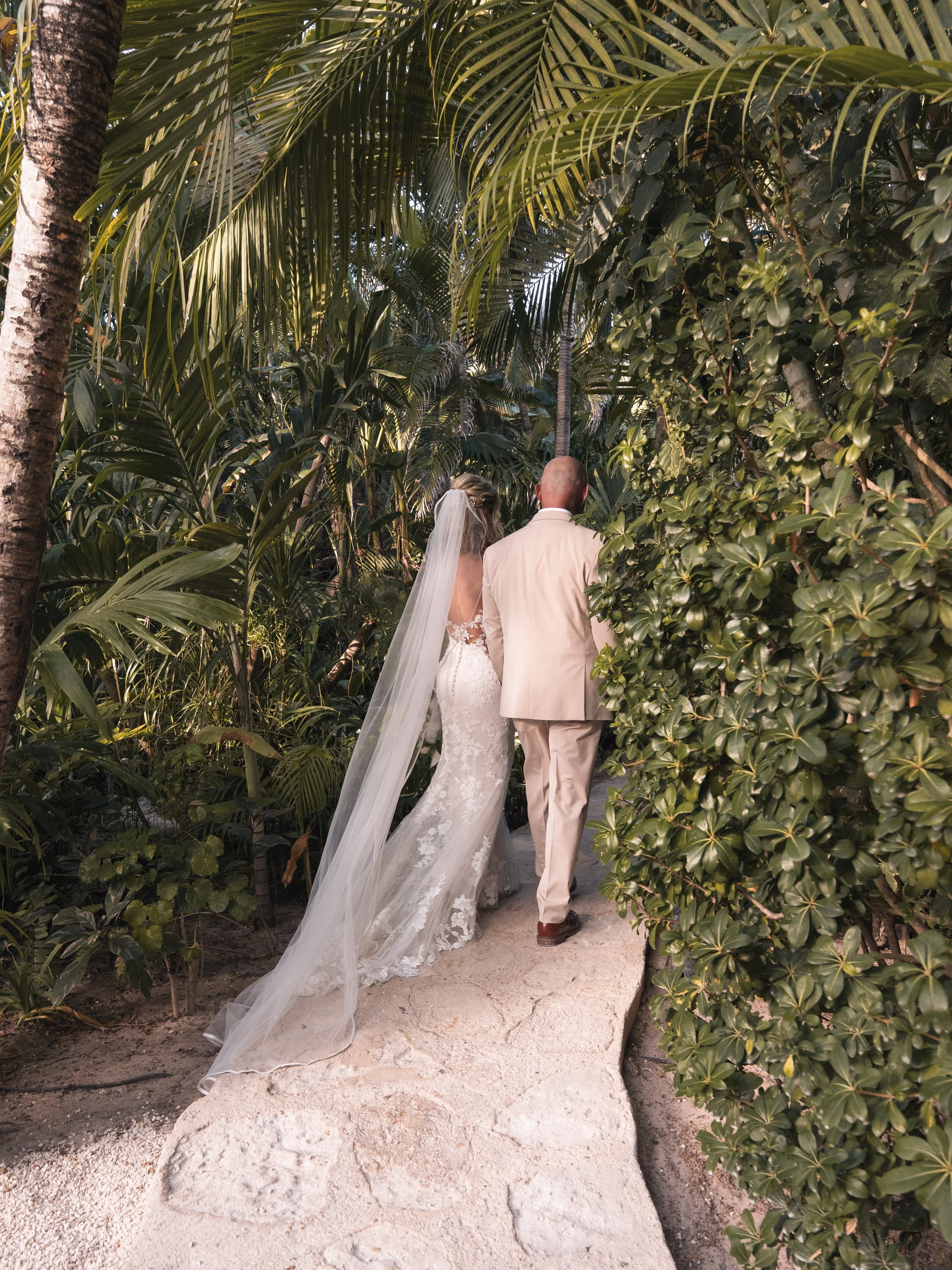 A bride and groom walking on a sandy pathway through a lush tropical garden with dense green foliage and tall palm trees, seen from behind.