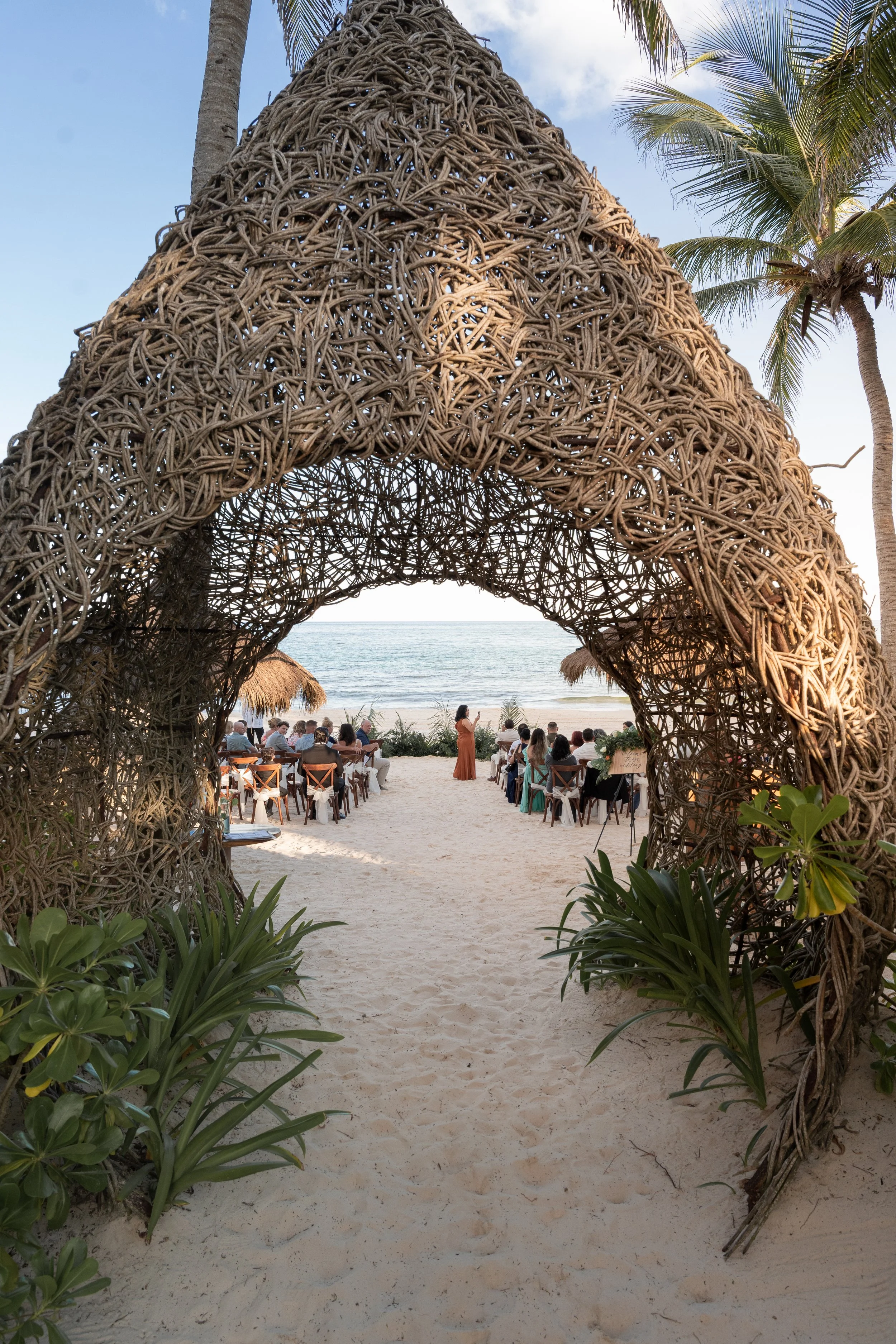 A beach wedding ceremony setup with chairs, a thatched arch, and guests, as seen through a wicker archway, near the ocean with palm trees and a cloudy sky.