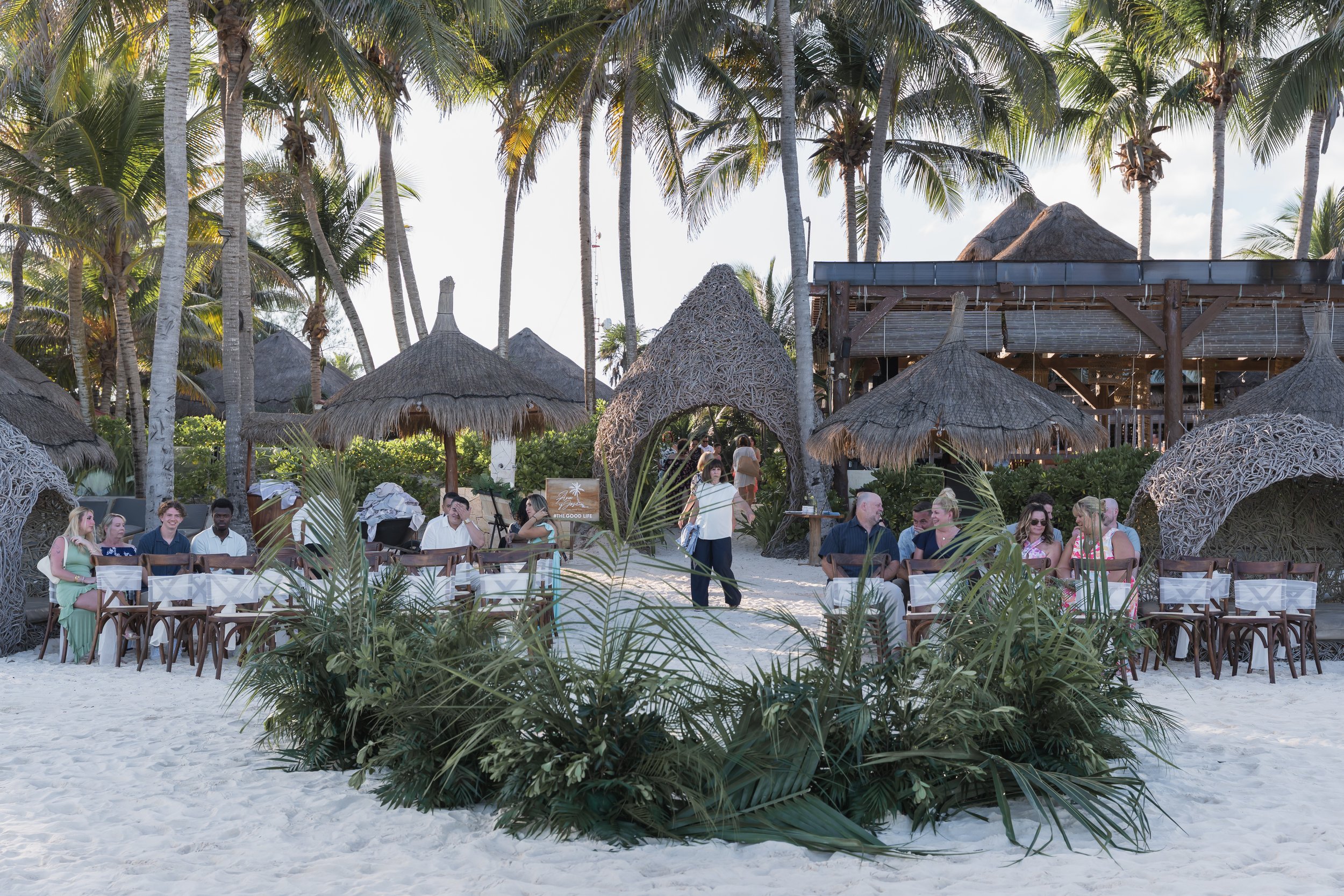 People dining outdoors on a beach at a tropical resort with palm trees and thatched umbrellas.