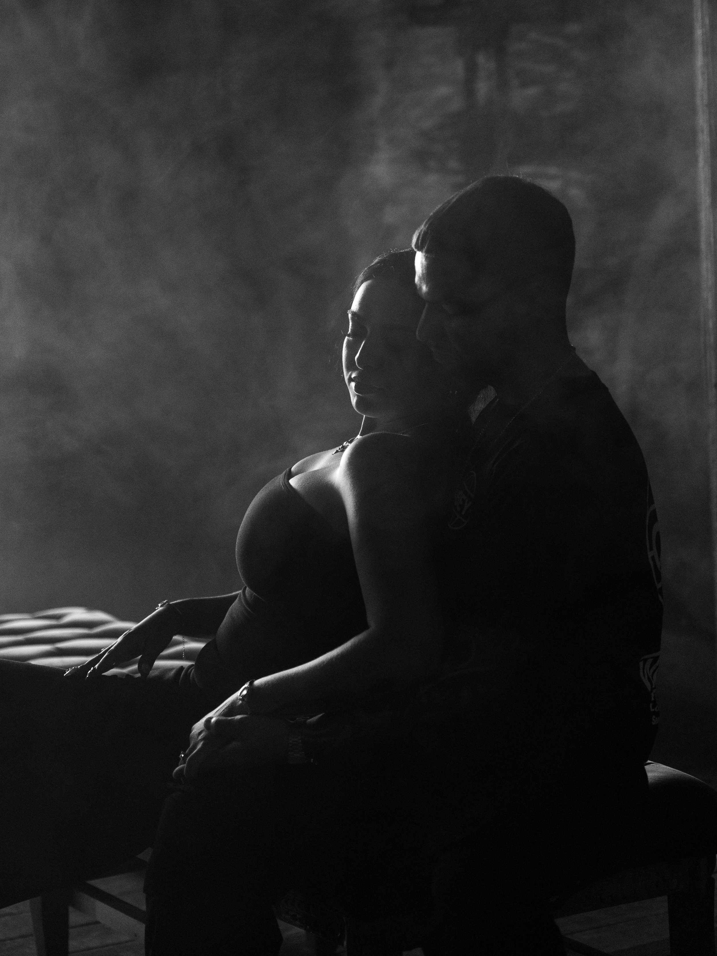 Silhouette of a couple sitting close together in a dimly lit room with a textured wall background, creating a romantic or intimate atmosphere.