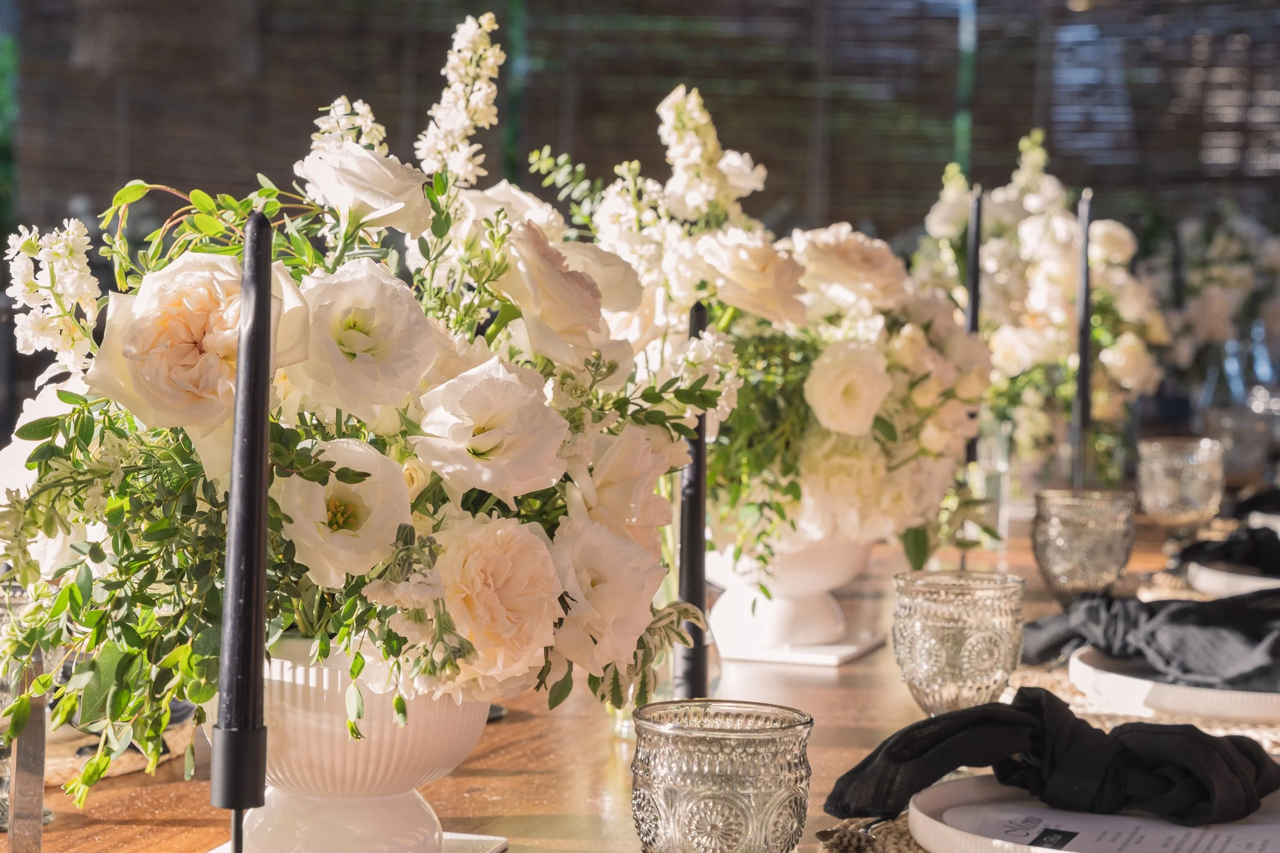 A table set with white floral centerpieces, clear textured glass cups, black napkins, and white plates, outdoors with a wooden surface.