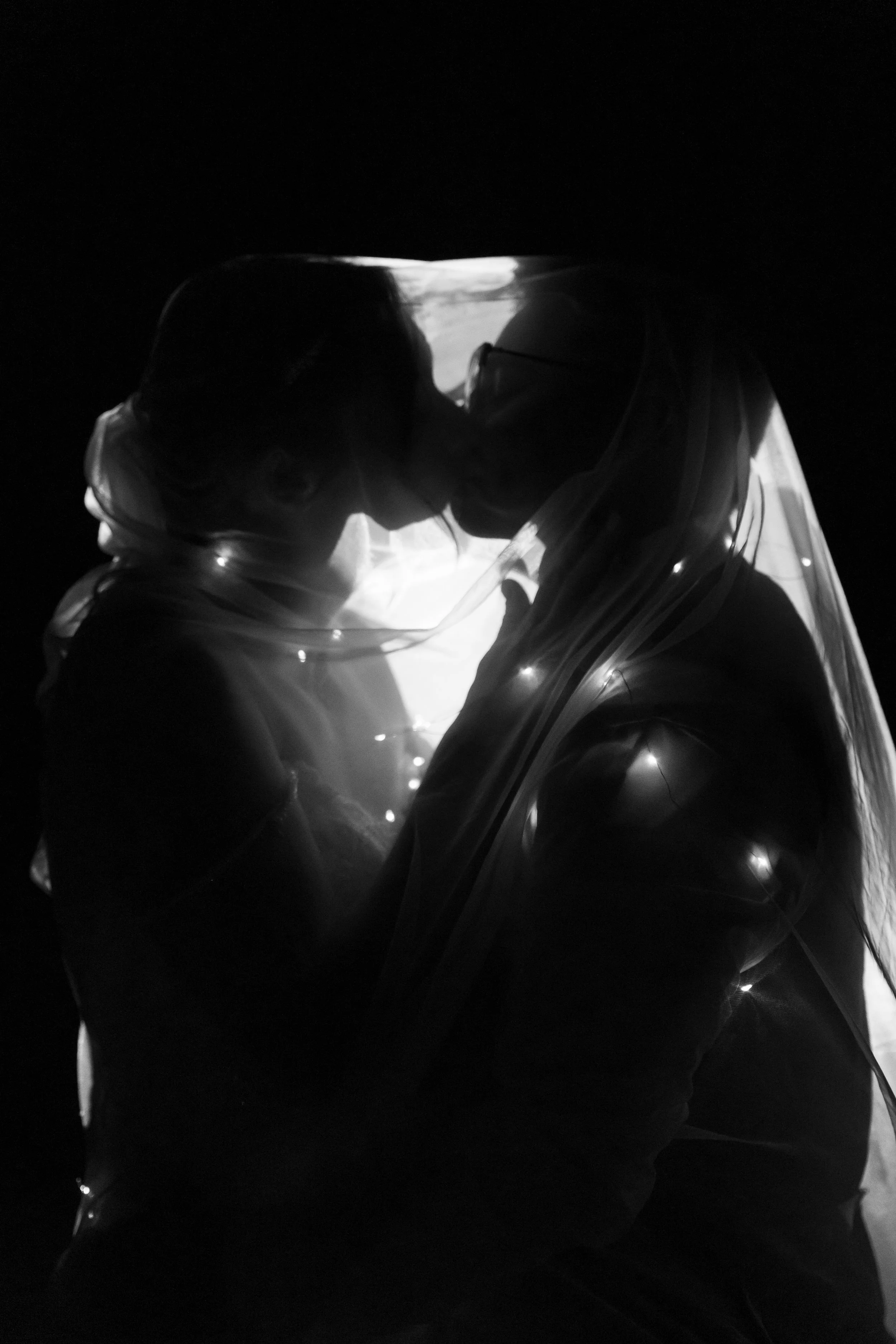 Silhouetted couple wearing face masks and a transparent plastic face shield, sharing a kiss in a dark setting with soft light.